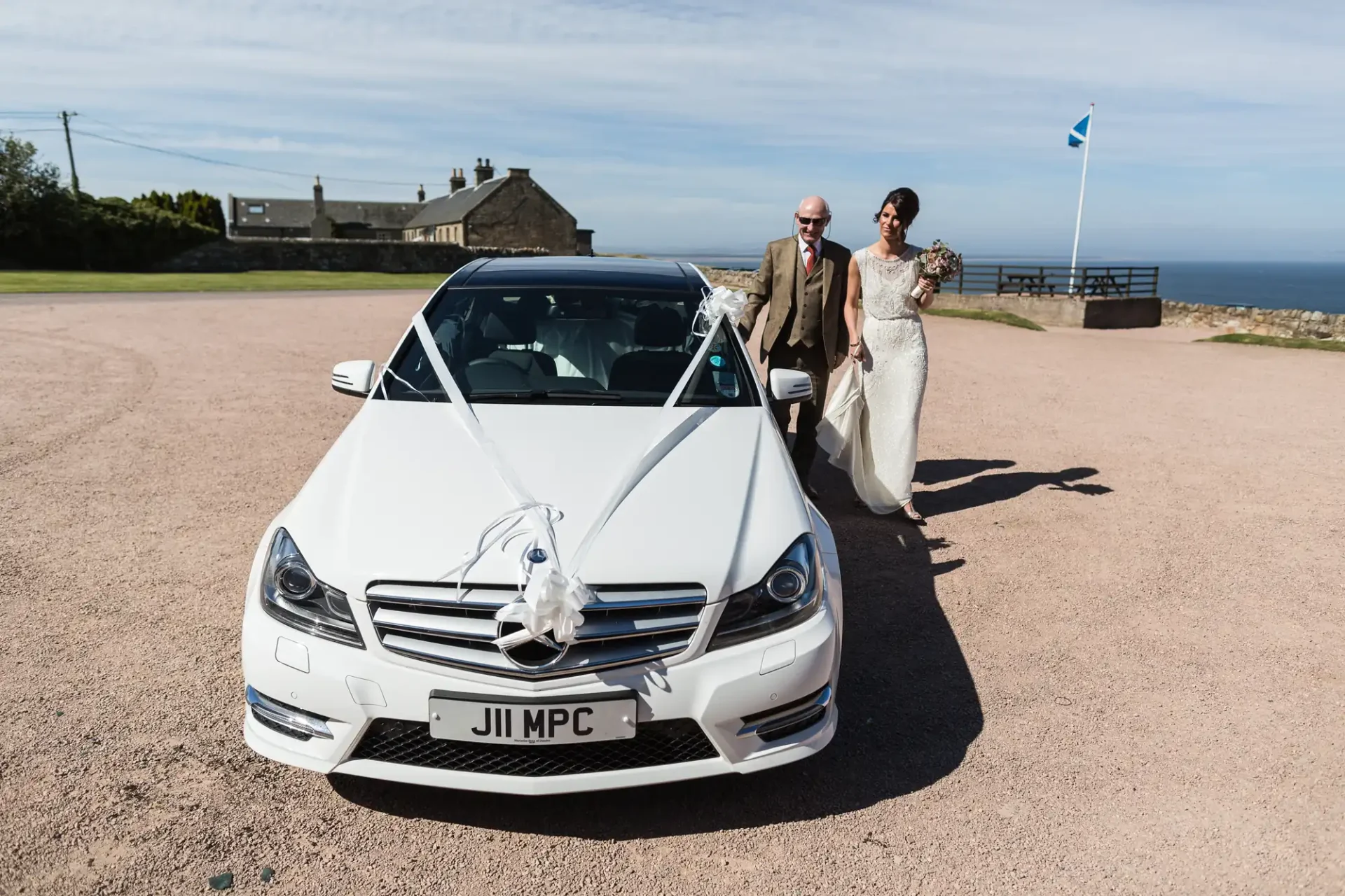 A bride and groom walking hand in hand beside a white decorated convertible car, with a scenic coastal backdrop and a clear blue sky.
