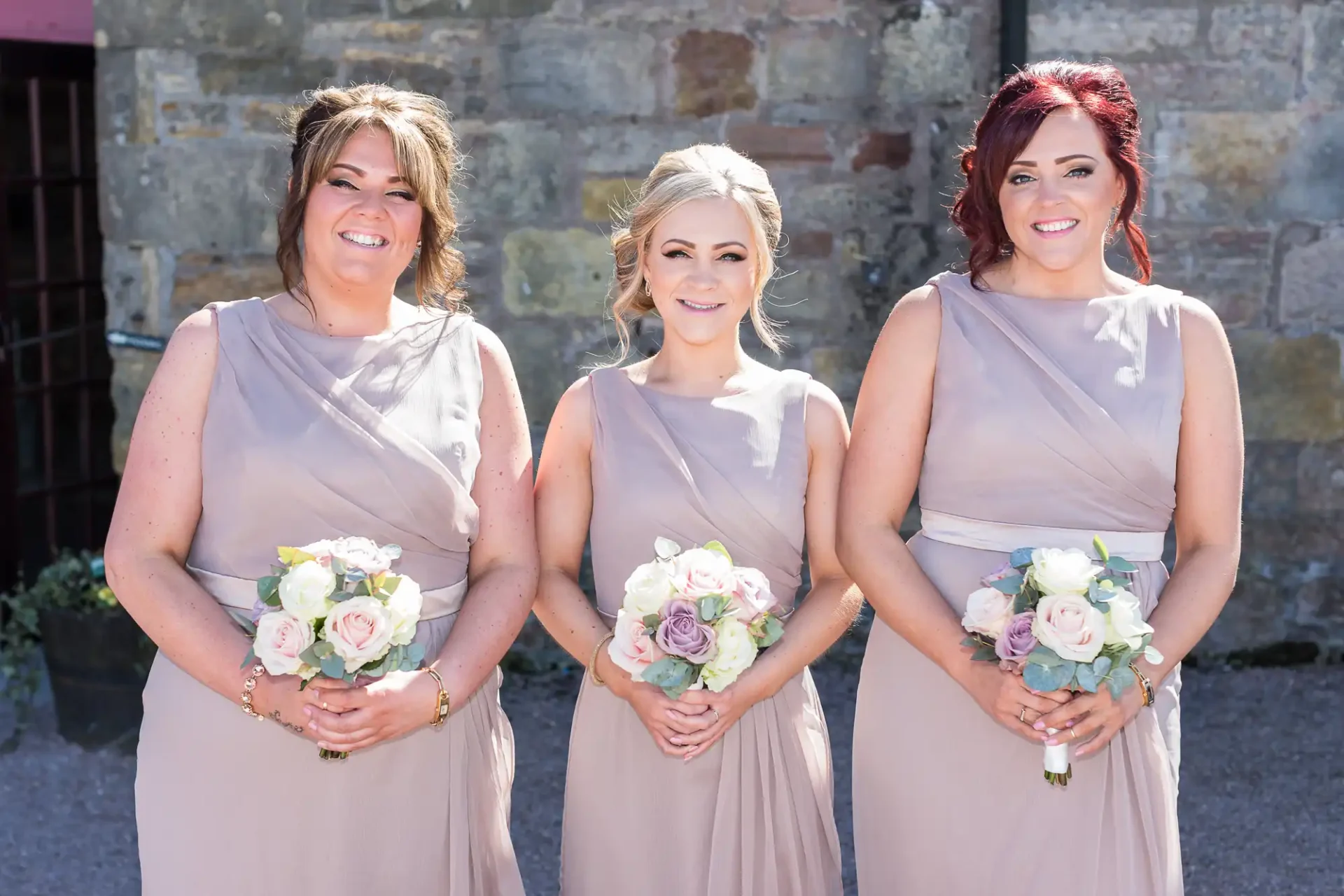 Three bridesmaids in matching taupe dresses holding bouquets of pink and white roses stand smiling outside a stone building.