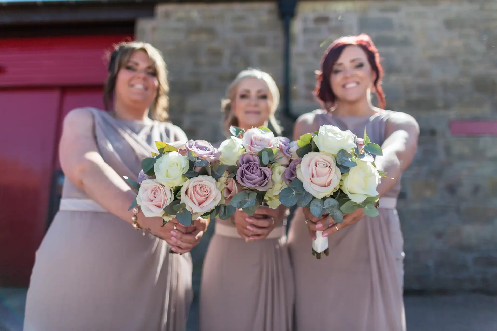 Three bridesmaids in taupe dresses holding bouquets of pink and purple roses, smiling, with a blurred building background.