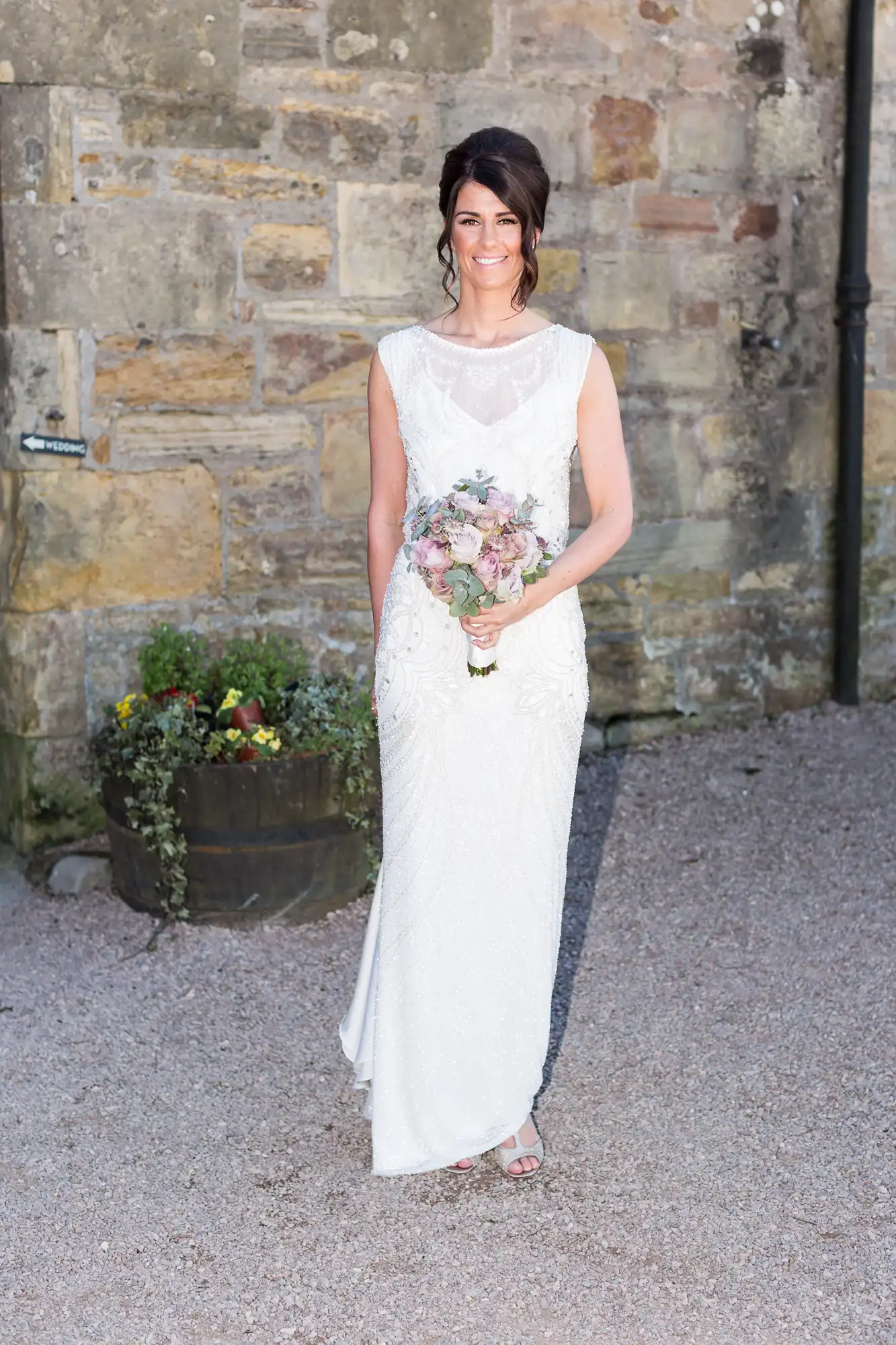 A bride in a white sleeveless gown smiling with a bouquet of flowers, standing outside in front of a stone building.