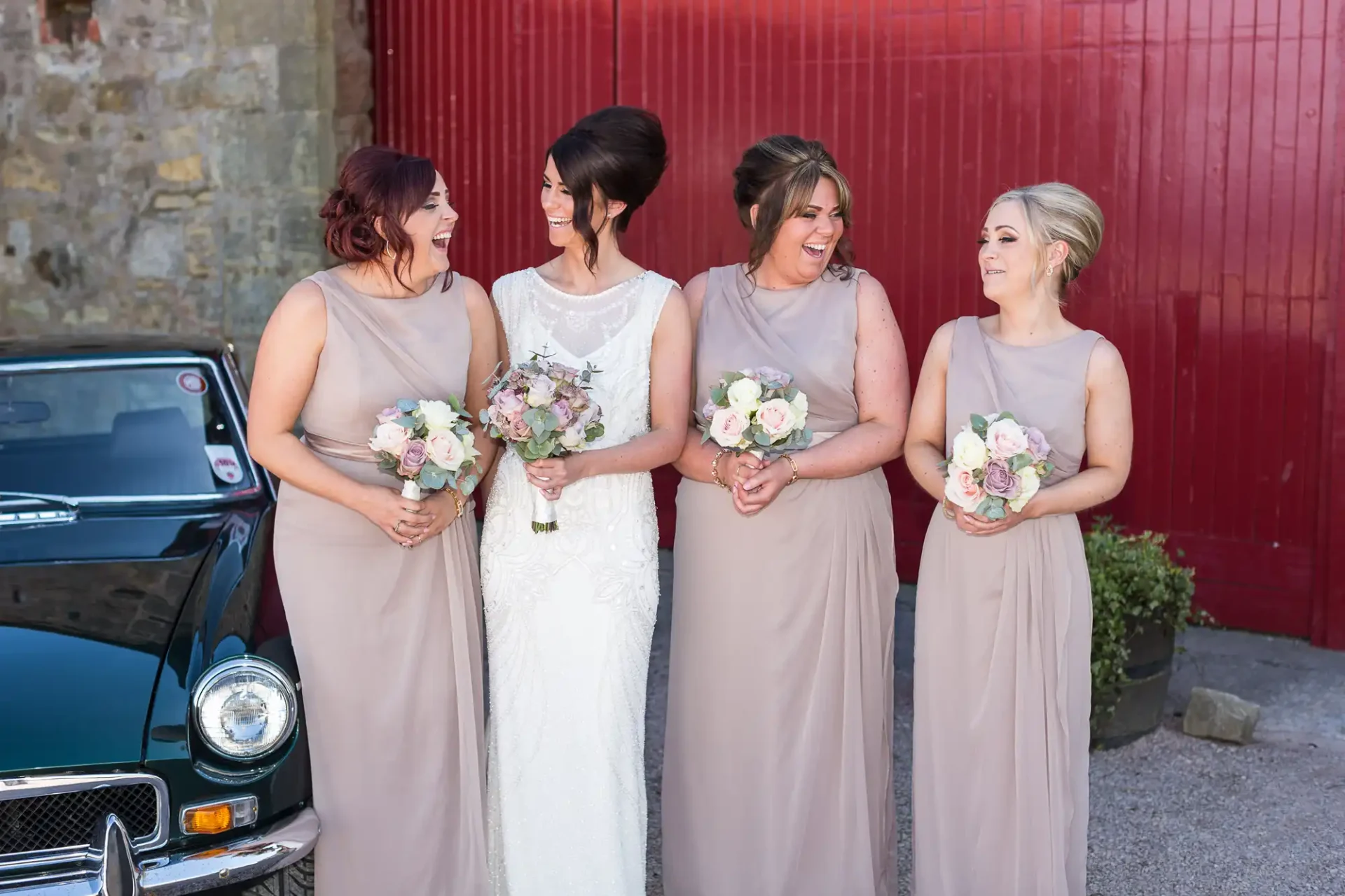 A bride and three bridesmaids in beige dresses laughing together in front of a vintage car and a red door.