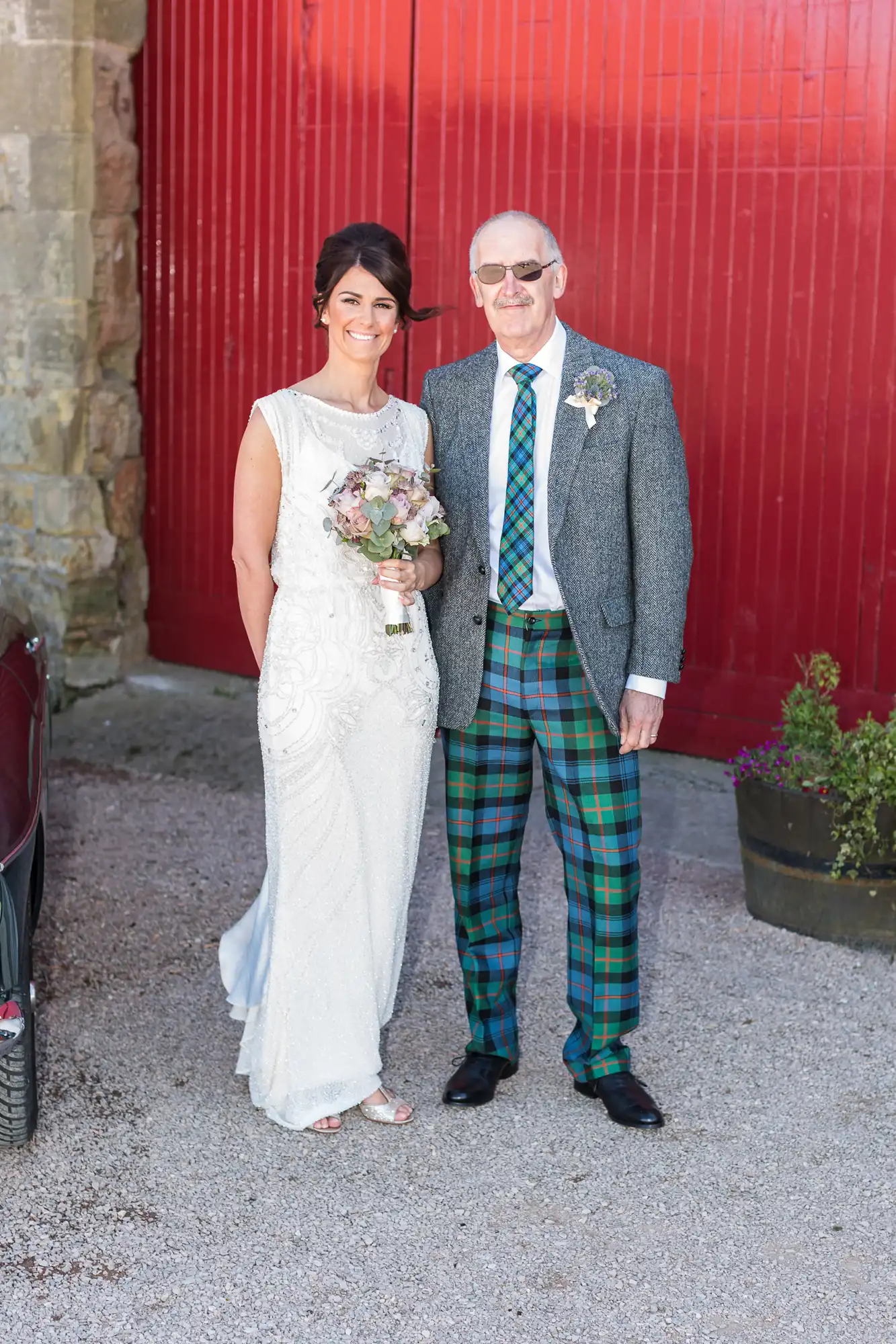 A bride in a white beaded gown stands beside a groom in a grey jacket and vibrant tartan trousers, both smiling, against a red barn backdrop.