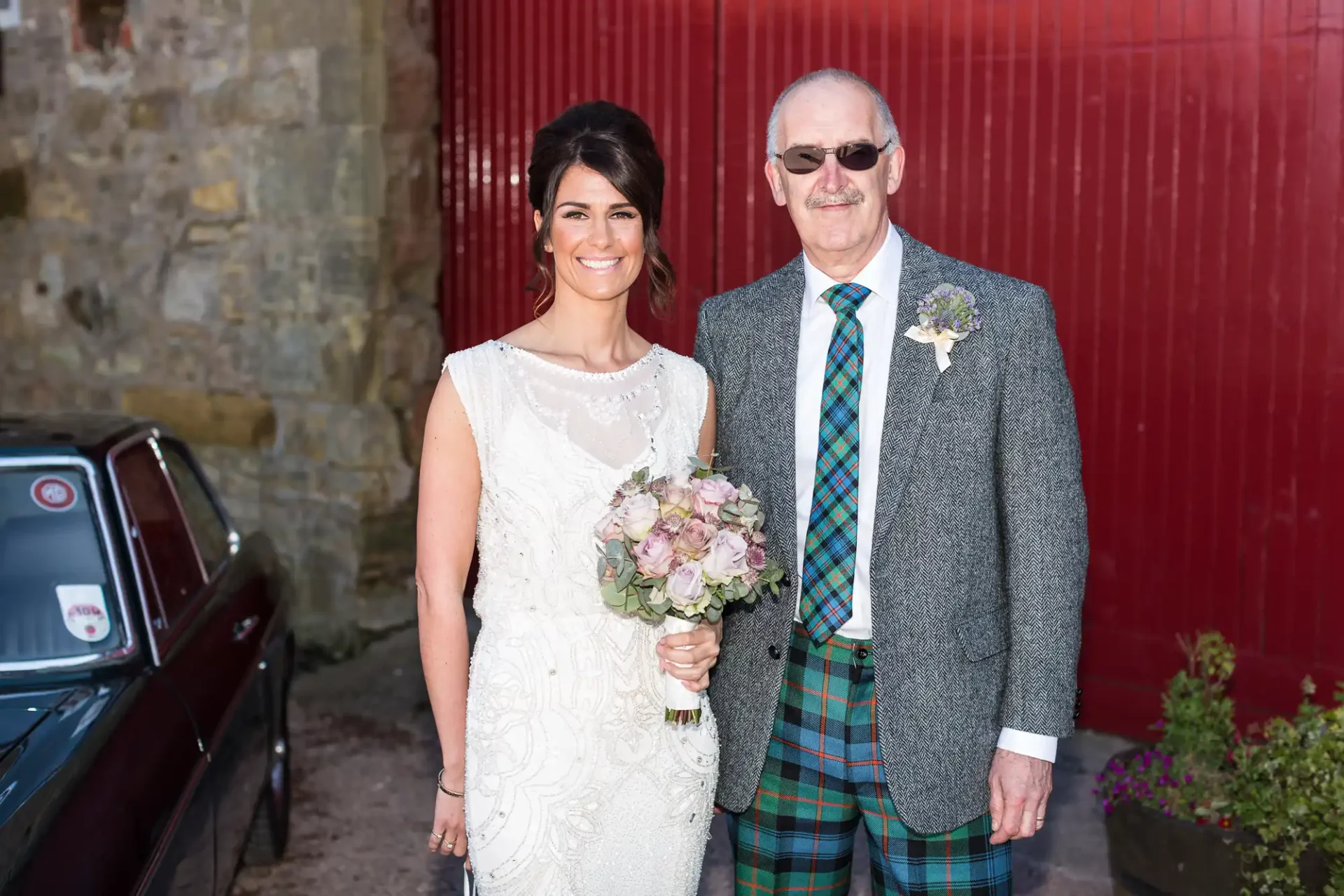A bride in a white dress and a groom in a tartan kilt and gray jacket pose smiling beside a vintage car against a red background.