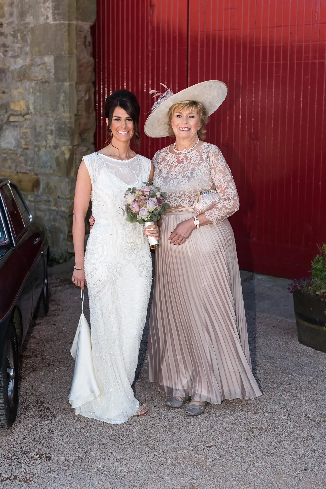 Two women dressed in formal wear posing with a bouquet, one in a wedding dress and the other in a long dress and hat, beside a vintage car.