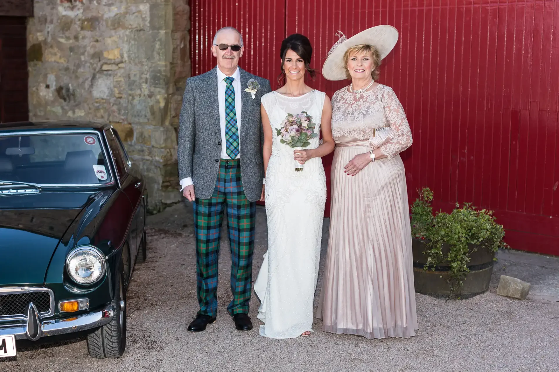 A bride in a white dress stands with an older couple by a classic car; the man in a plaid kilt and the woman in a beige dress and hat.