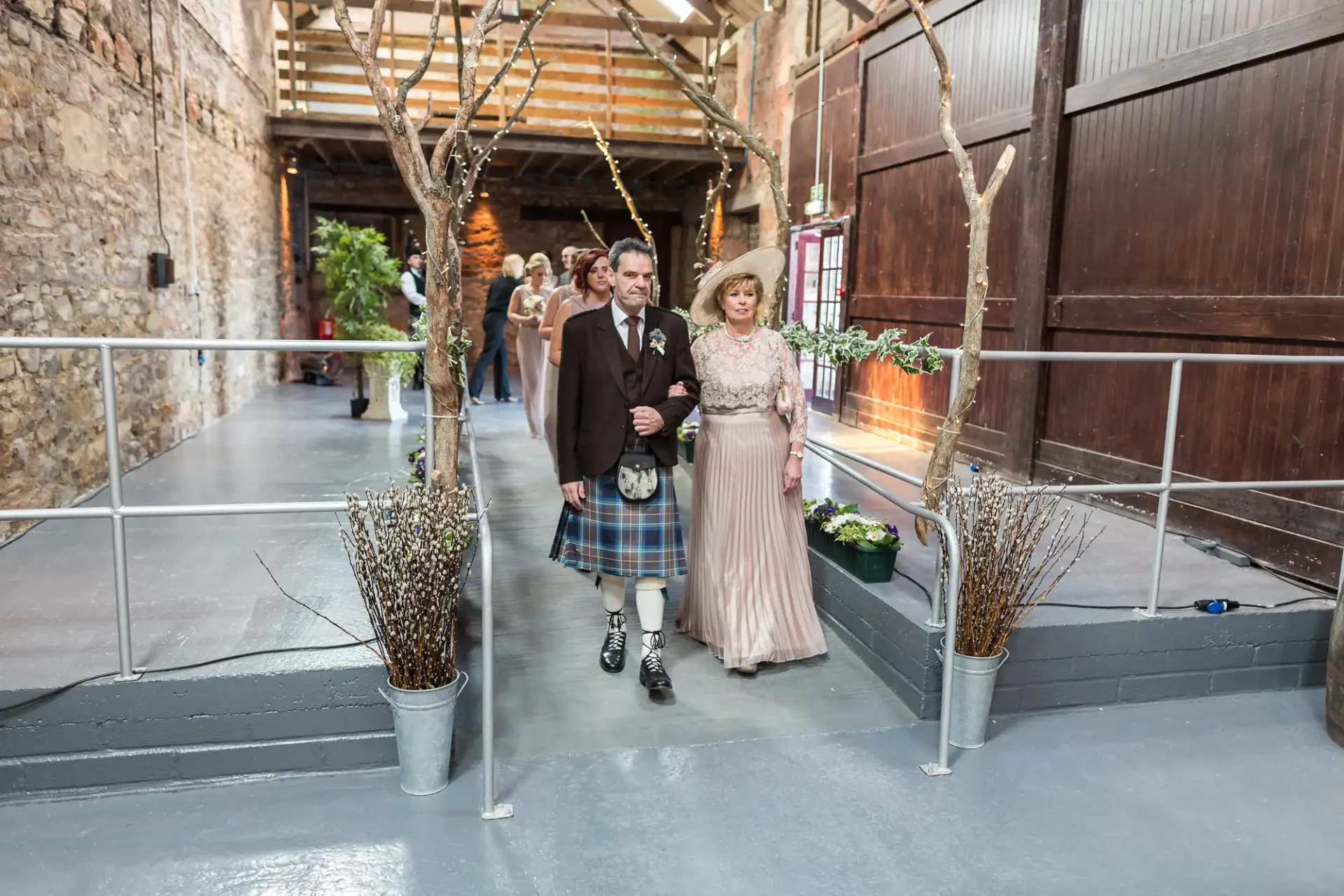 A couple dressed in traditional scottish attire walking down an aisle in a rustic indoor venue, flanked by decorative trees.