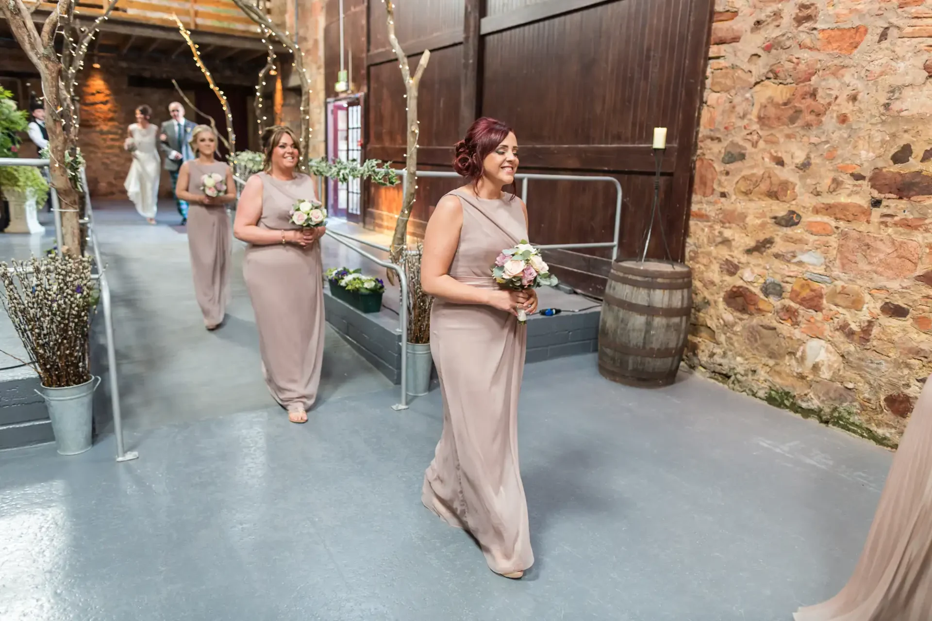 Three bridesmaids walk down an aisle in a rustic venue, holding bouquets, dressed in long beige dresses.