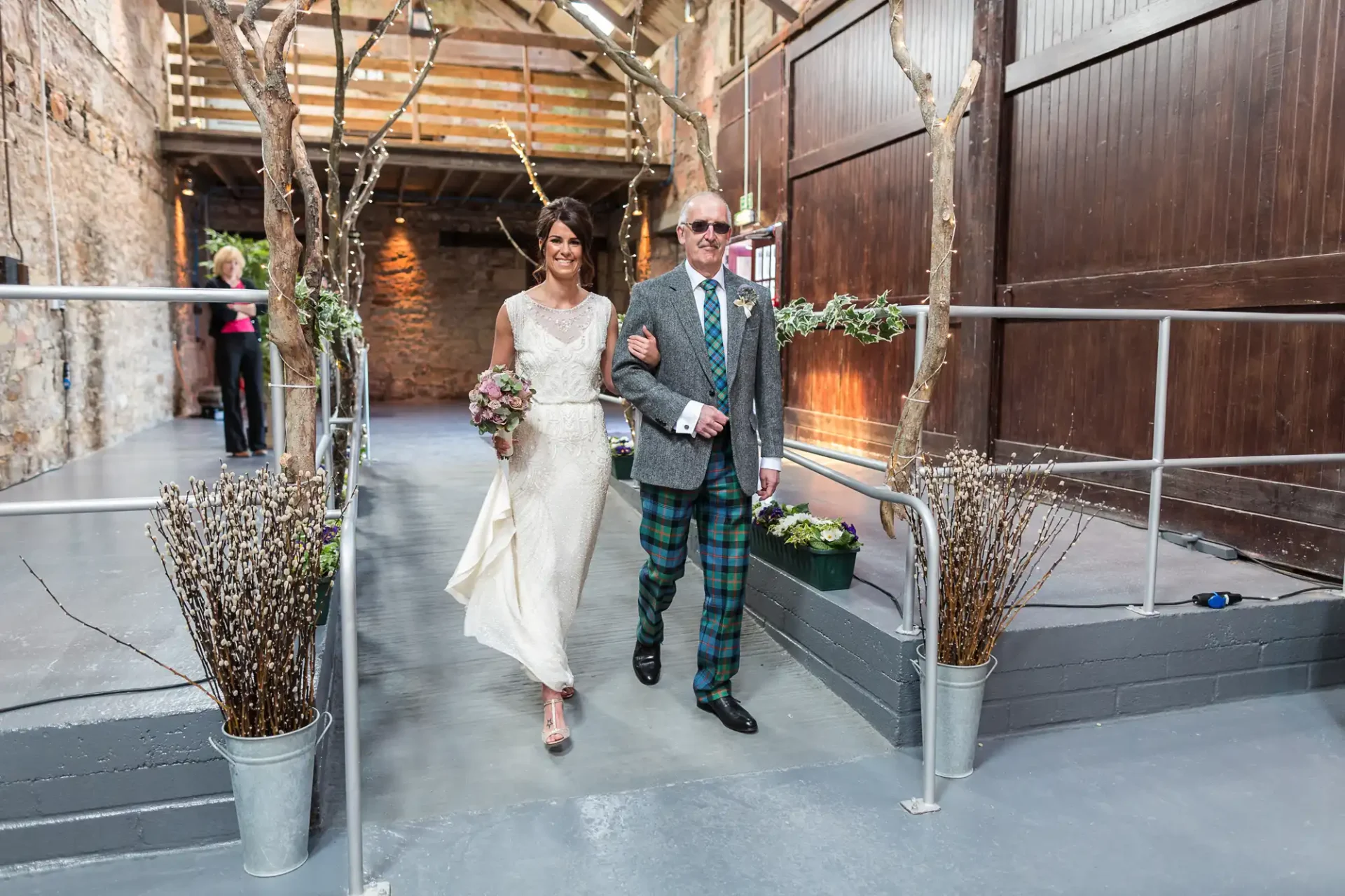 A bride and her father walking down the aisle in a rustic indoor setting, the father wearing a tartan kilt and the bride in a white lace dress.