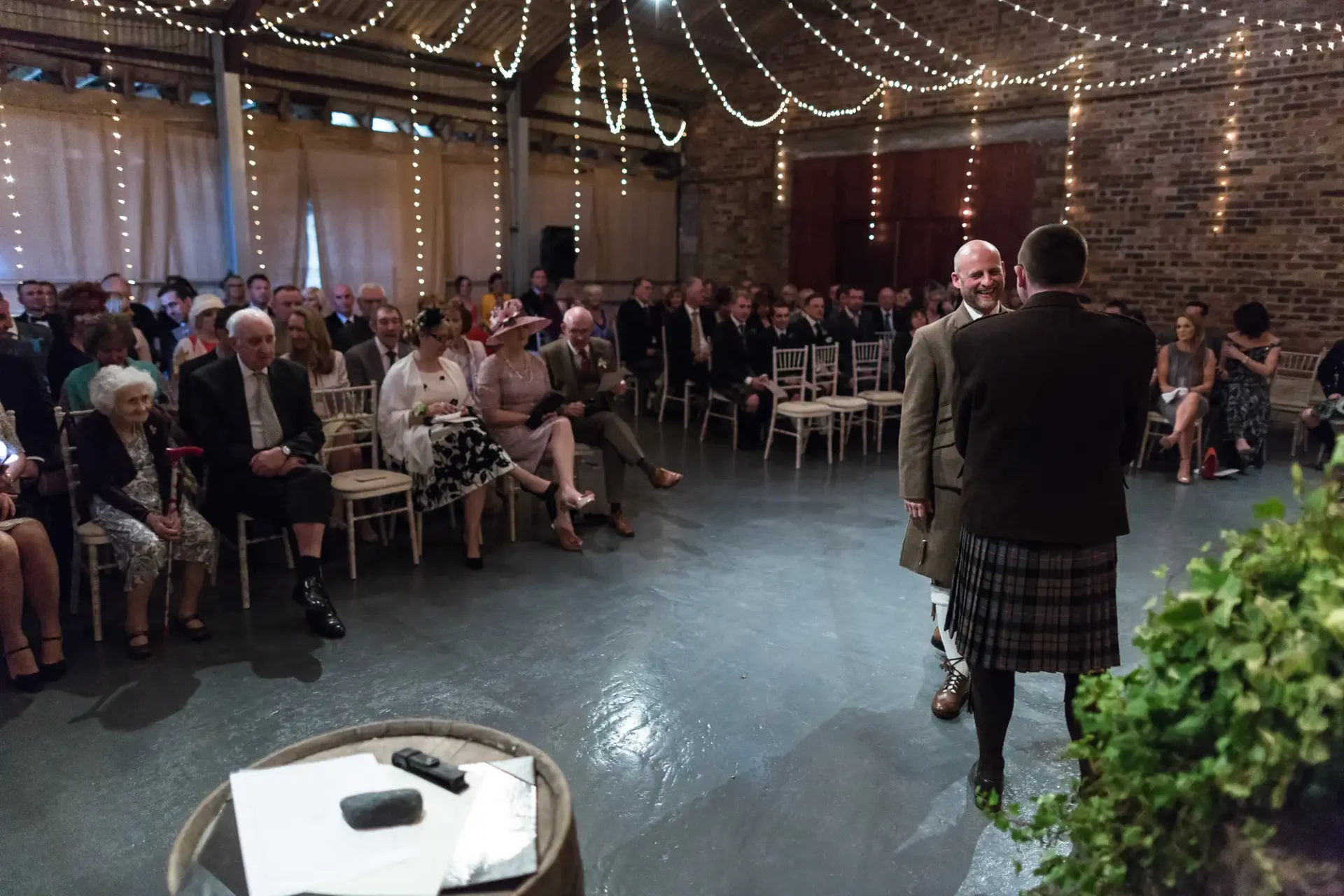 A wedding reception in a rustic venue with guests watching two men in suits, one wearing a kilt, smiling at each other near a microphone stand.
