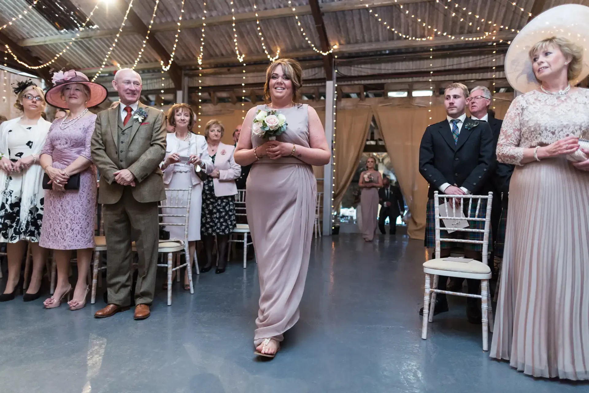 A bride walks down the aisle, smiling and holding a bouquet, as guests dressed in formal attire watch in a rustic, barn-like venue.