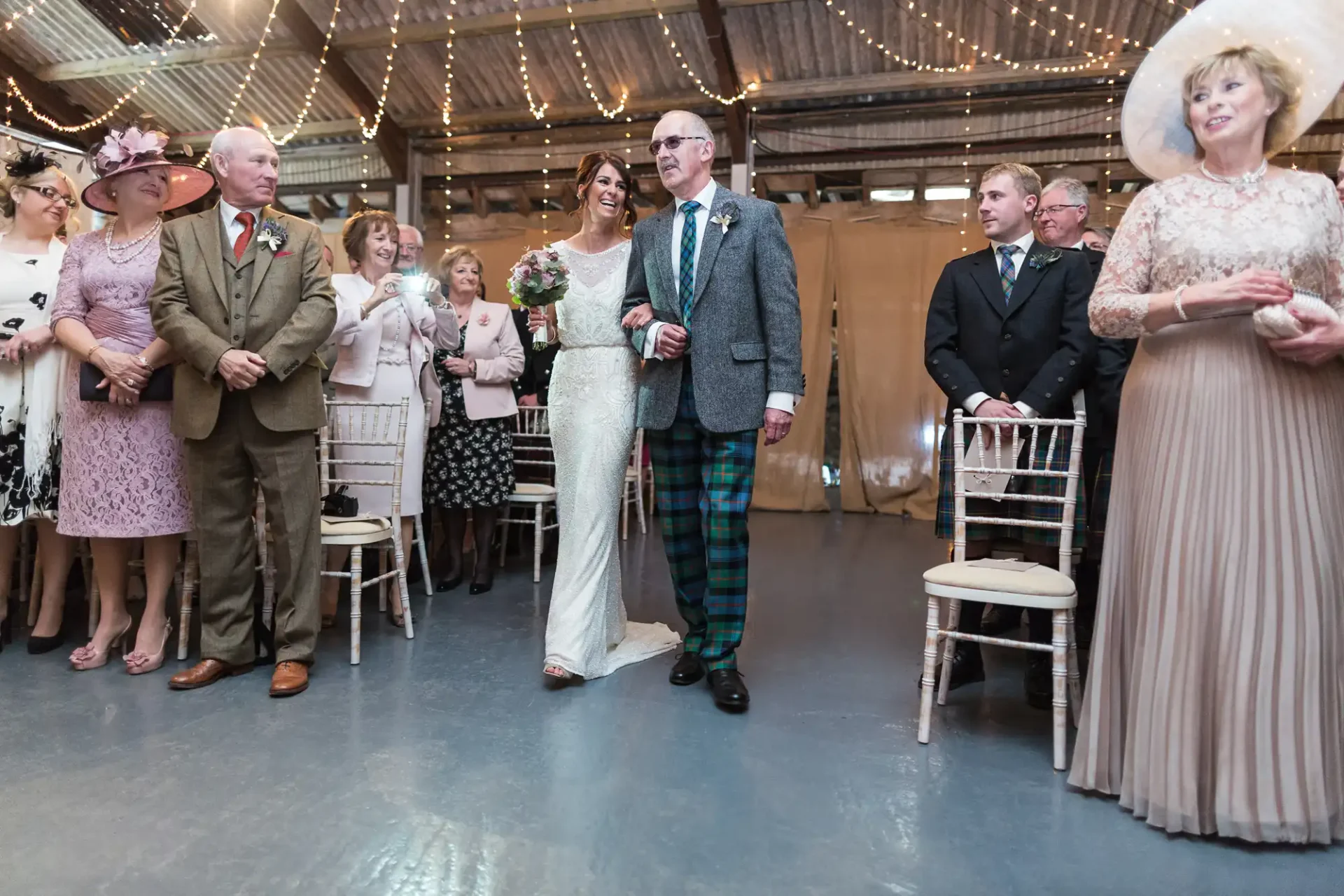 Bride and groom walking down the aisle, surrounded by guests in elegant attire, in a rustic indoor setting.