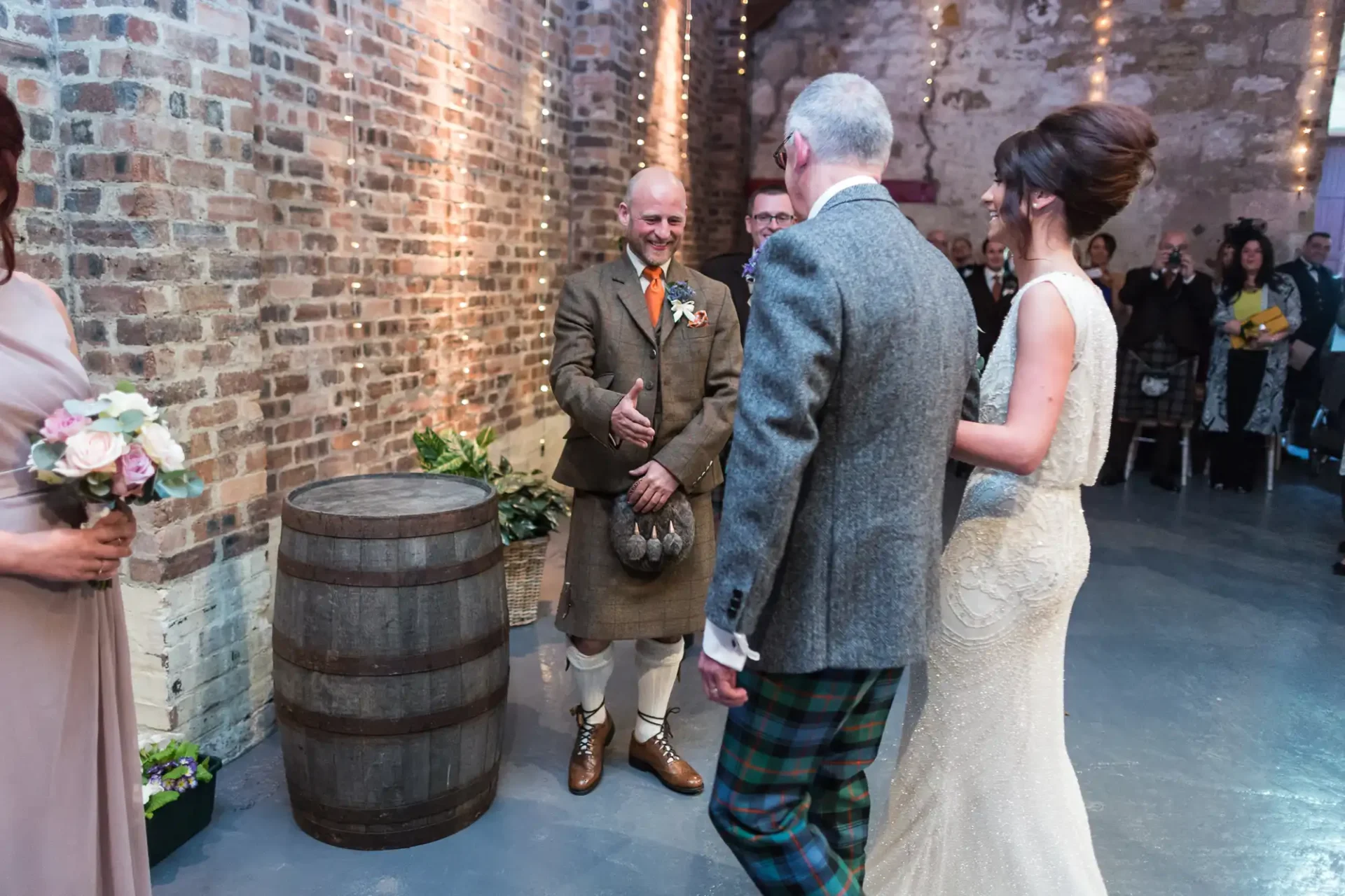 A wedding ceremony in a rustic venue with brick walls, featuring a smiling officiant and a couple holding hands, surrounded by guests.