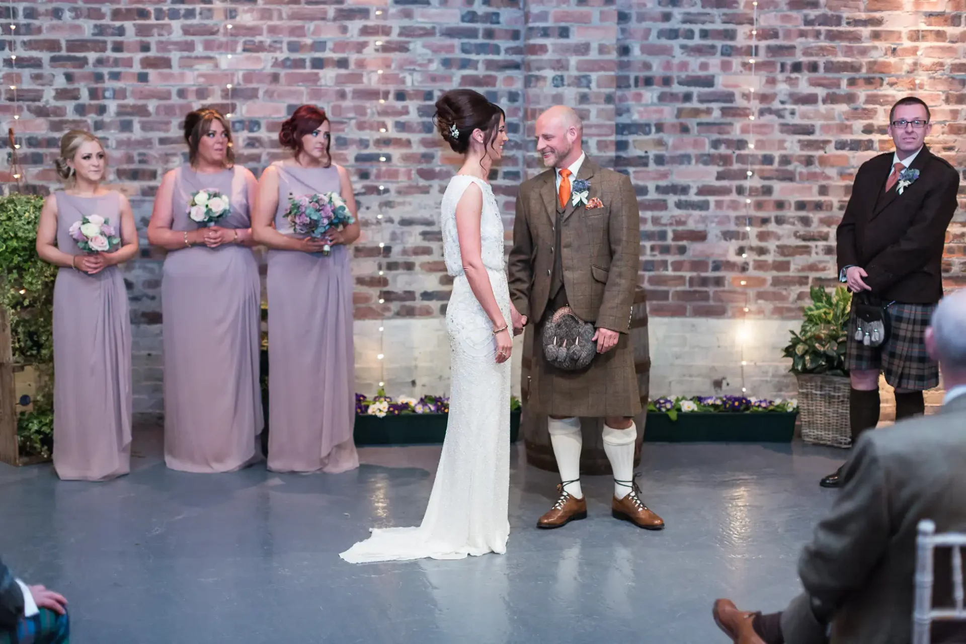 A bride and groom face each other smiling, surrounded by bridesmaids in pastel dresses and a best man in a kilt, in a venue with brick walls.