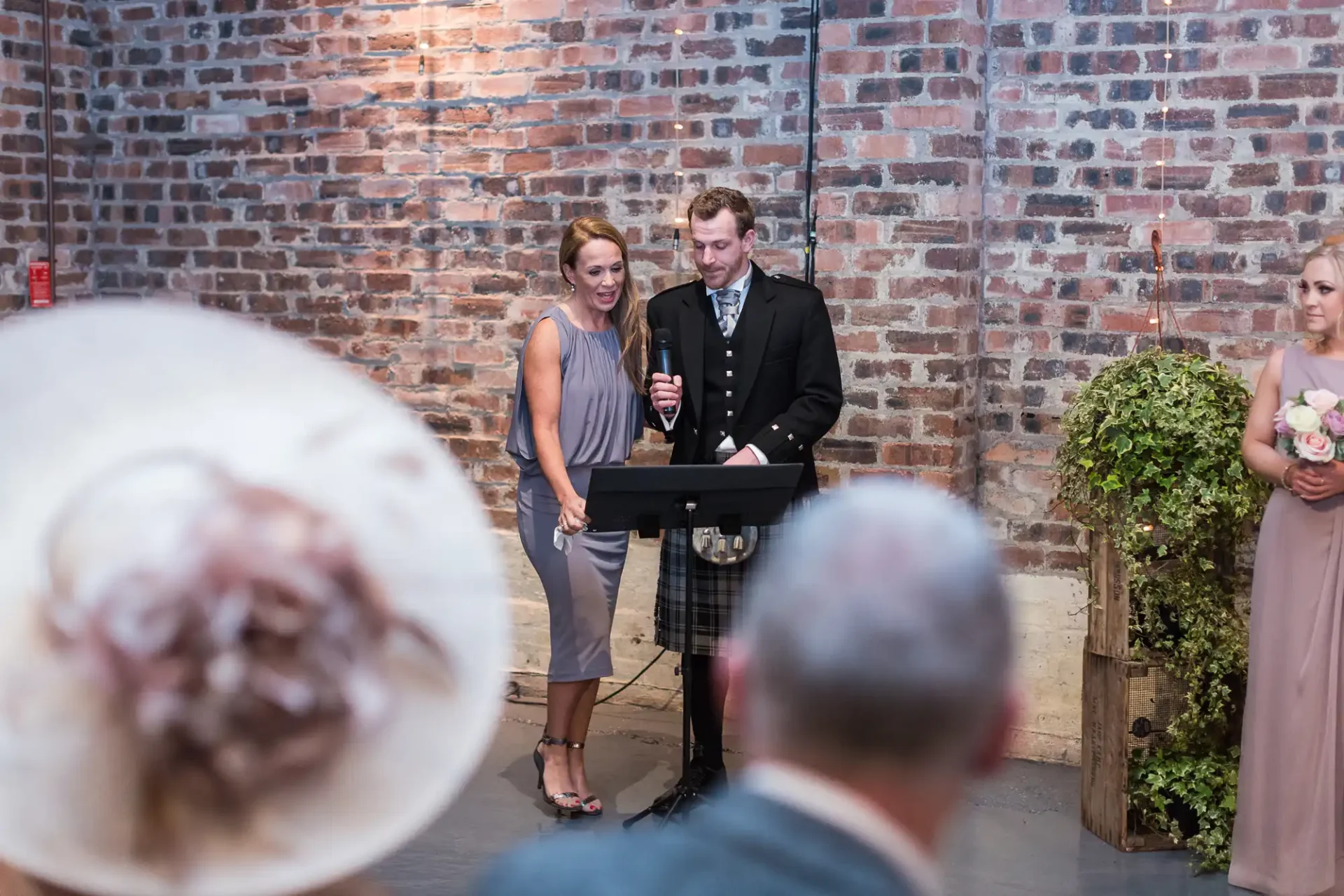 A man in a kilt and a woman holding a microphone deliver a speech at a wedding, with guests listening in a venue with brick walls.