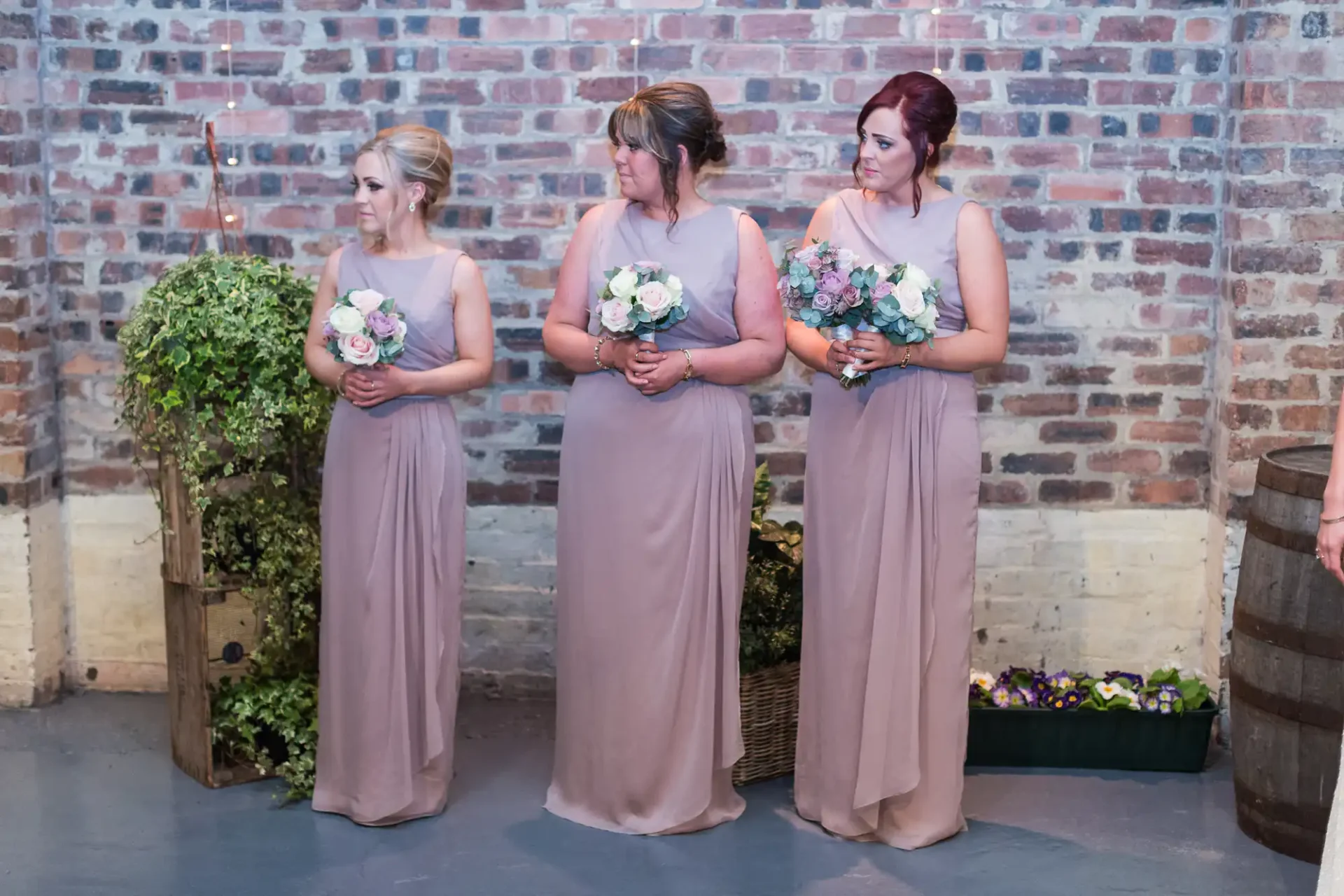 Three bridesmaids in matching pastel dresses holding bouquets, standing against a brick wall in a rustic setting.
