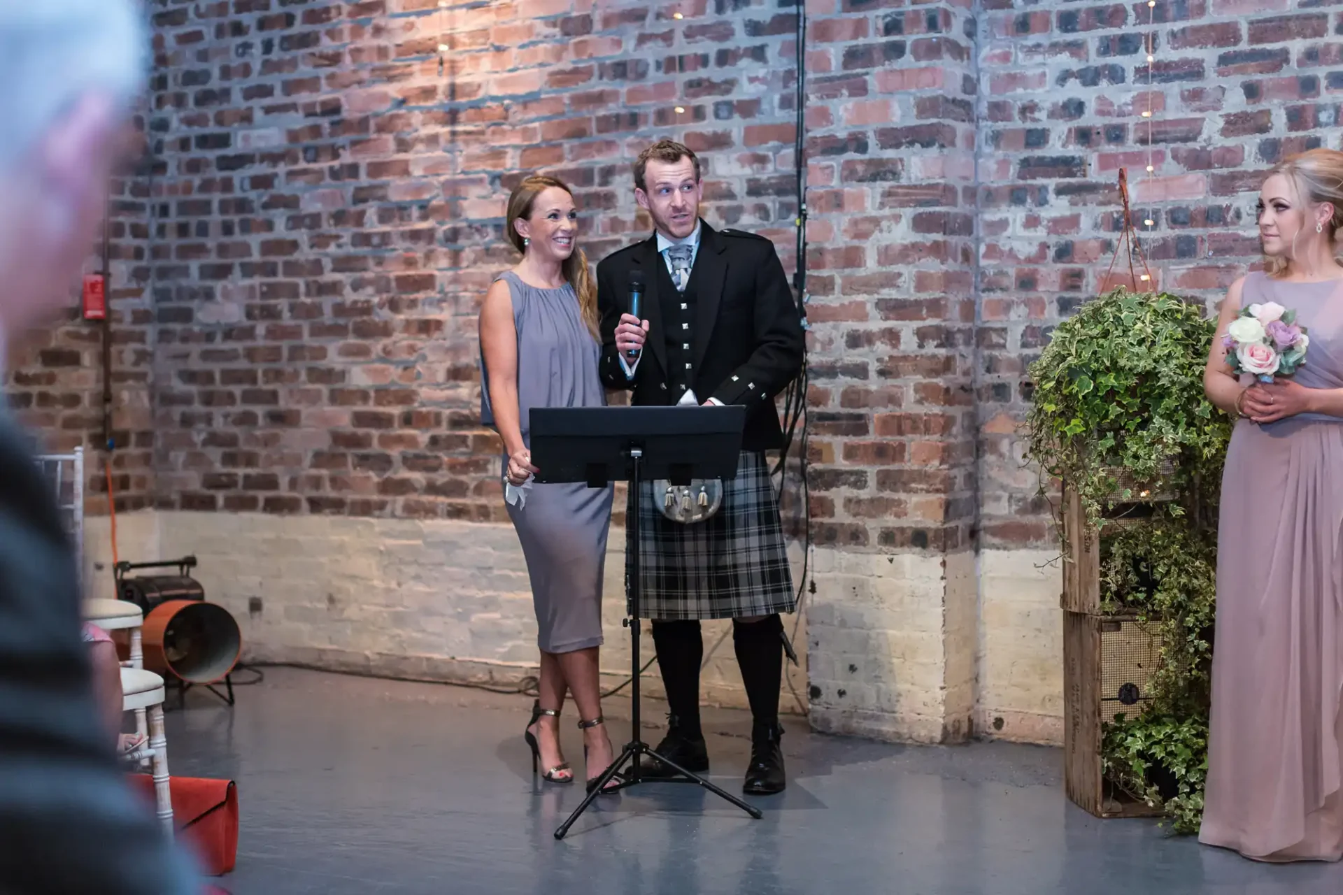 A man in a kilt and a woman in a gray dress speaking at a lectern during a wedding reception, with another woman holding flowers nearby. brick wall in the background.