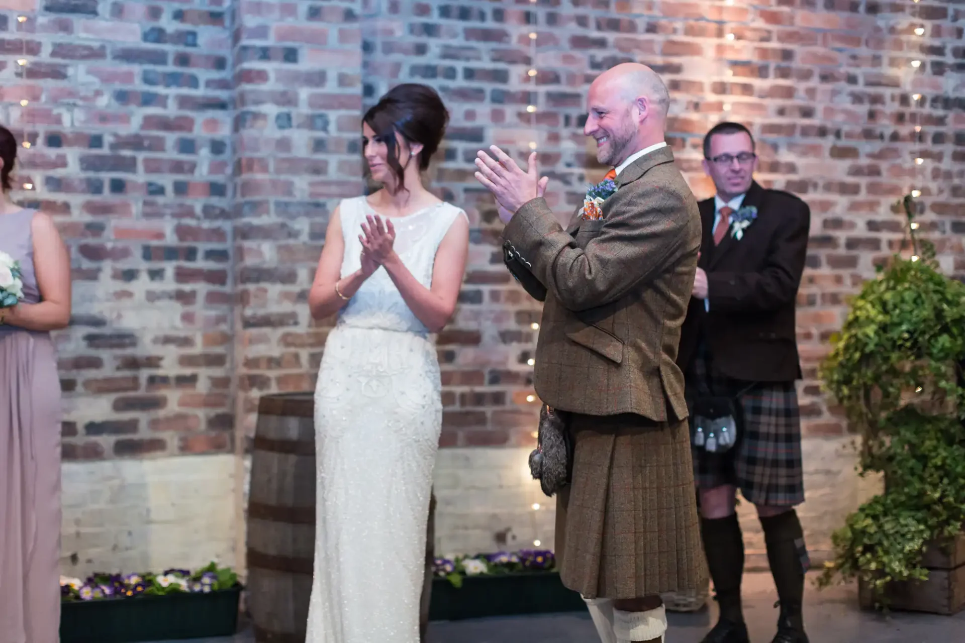 A bride and groom applauding, with the groom wearing a kilt, standing in front of a brick wall at an indoor wedding venue. a best man is visible in the background.
