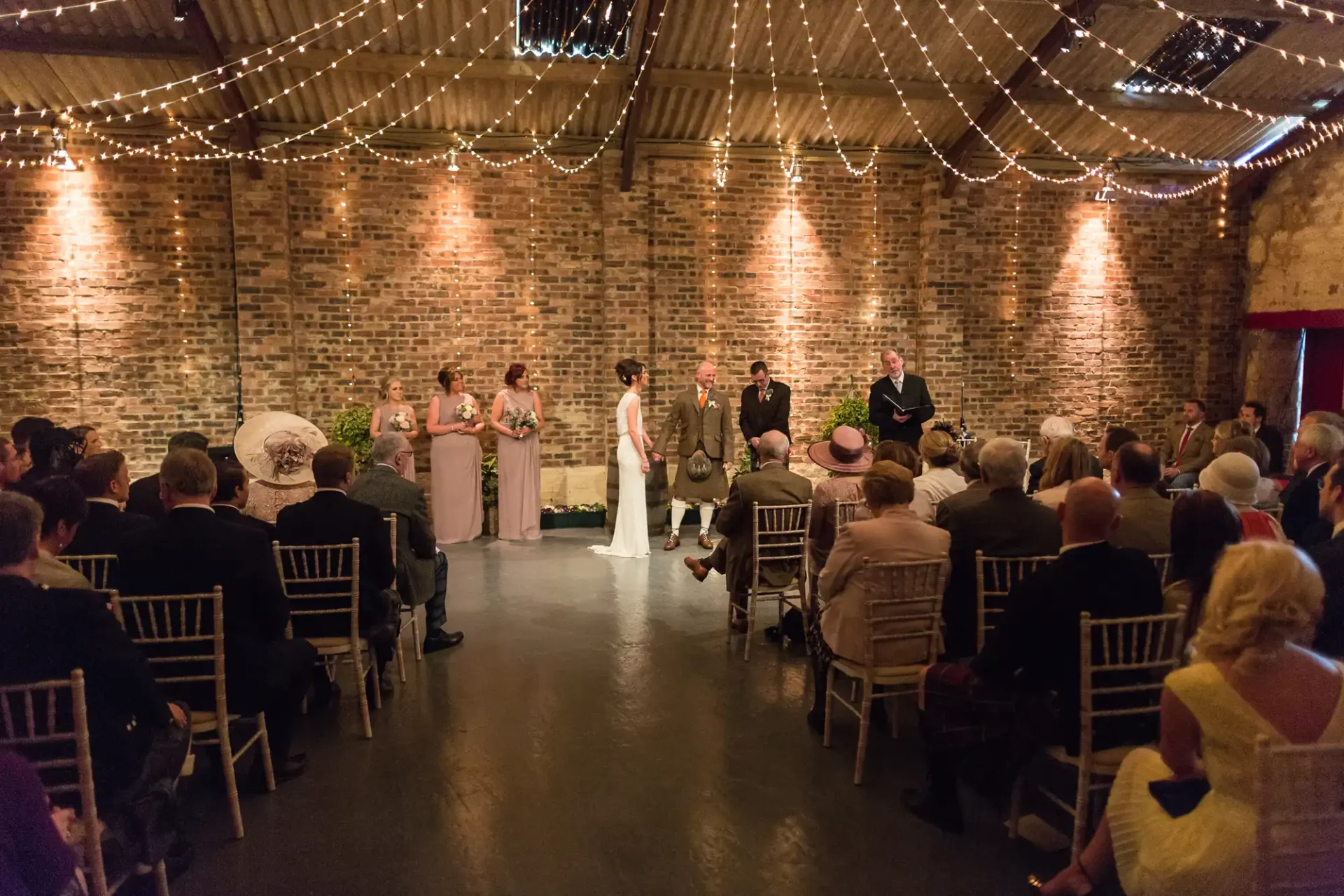 A wedding ceremony in a rustic venue with brick walls and string lights, featuring a couple at the altar, surrounded by guests.