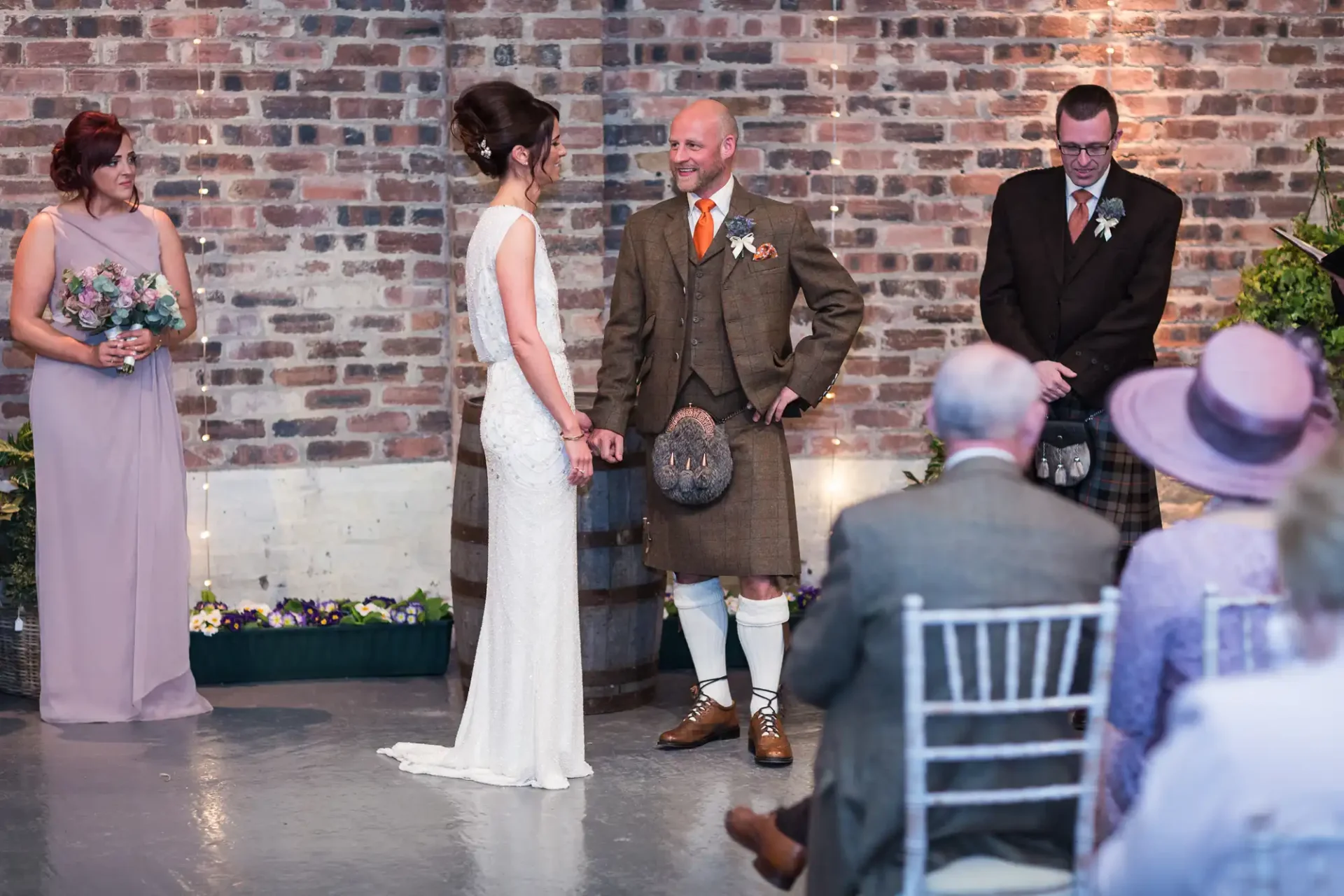 A bride and groom stand facing each other at their wedding, the groom in a kilt, with guests and a brick backdrop.