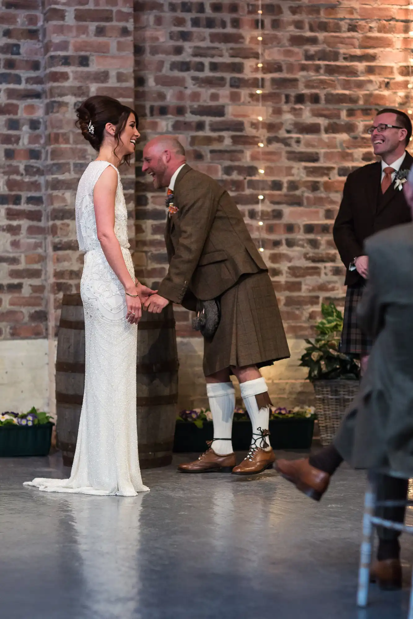 Bride in a white dress smiling at a groom in a kilt during a wedding ceremony, with a brick wall background and a guest watching.