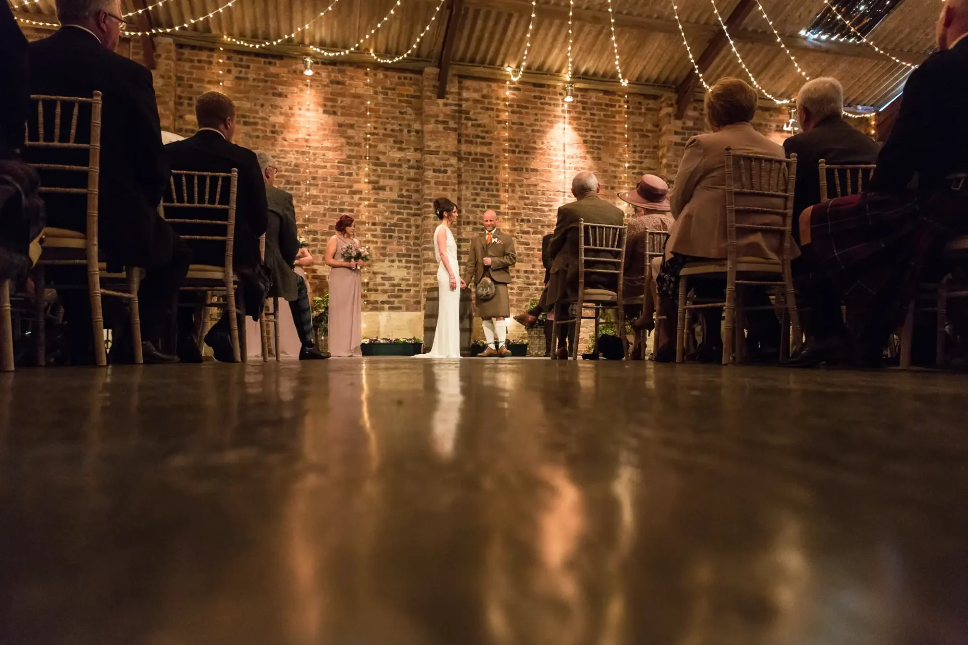A couple stands in front of an officiant during a wedding ceremony in a brick-walled venue, viewed from the floor level among seated guests.
