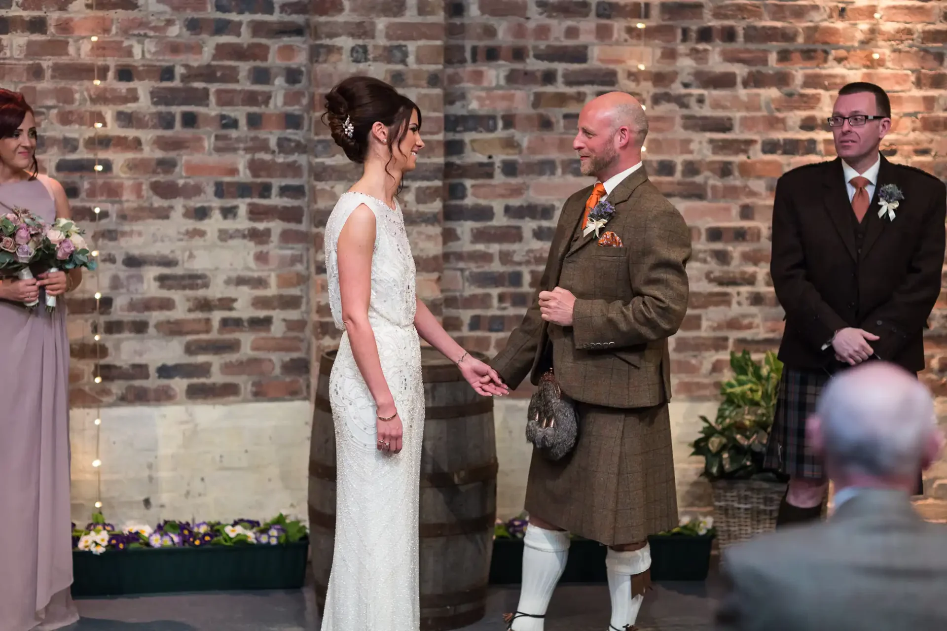 Bride and groom holding hands in wedding attire, groom in kilt, with witnesses nearby in a venue with brick walls.