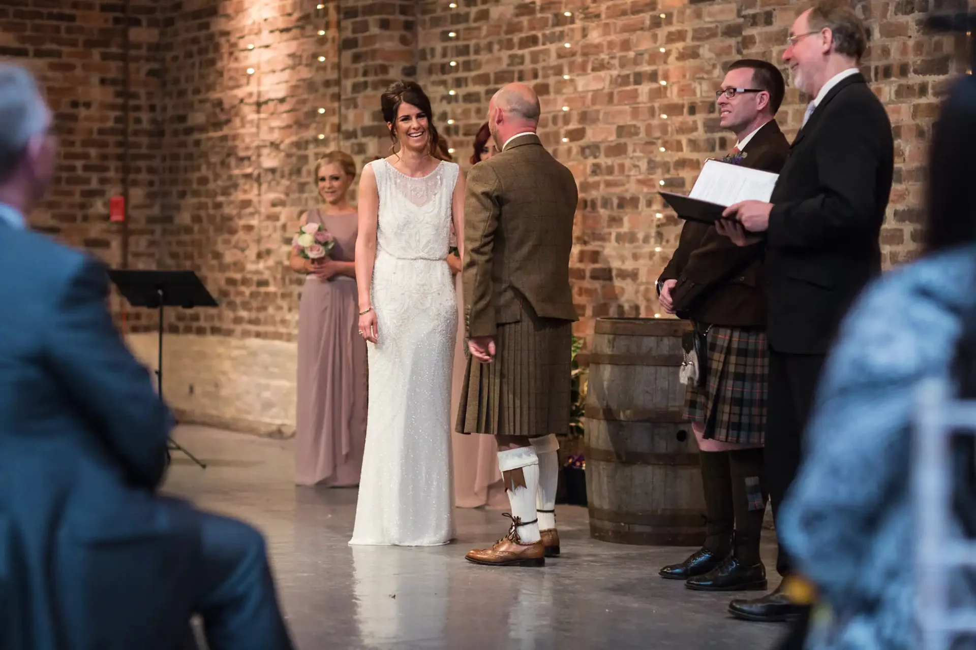 A bride in a white dress smiles during a wedding ceremony in a brick-walled venue with guests and a groom in a kilt.