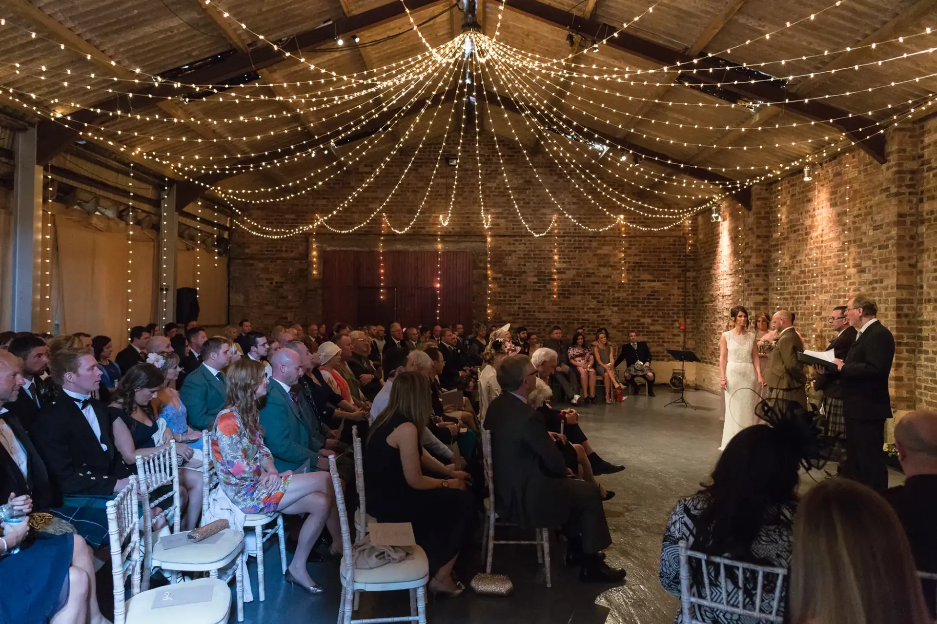 A wedding ceremony in a rustic brick hall, illuminated by strands of fairy lights, with guests seated watching the couple exchange vows.