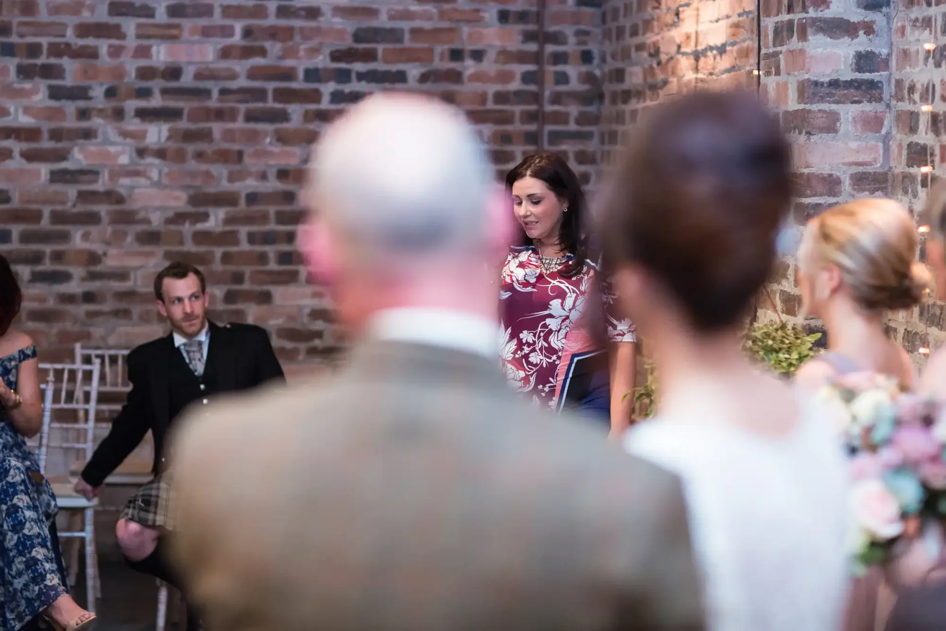 A woman speaks at a wedding, partially obscured by guests, in a room with brick walls and soft lighting.
