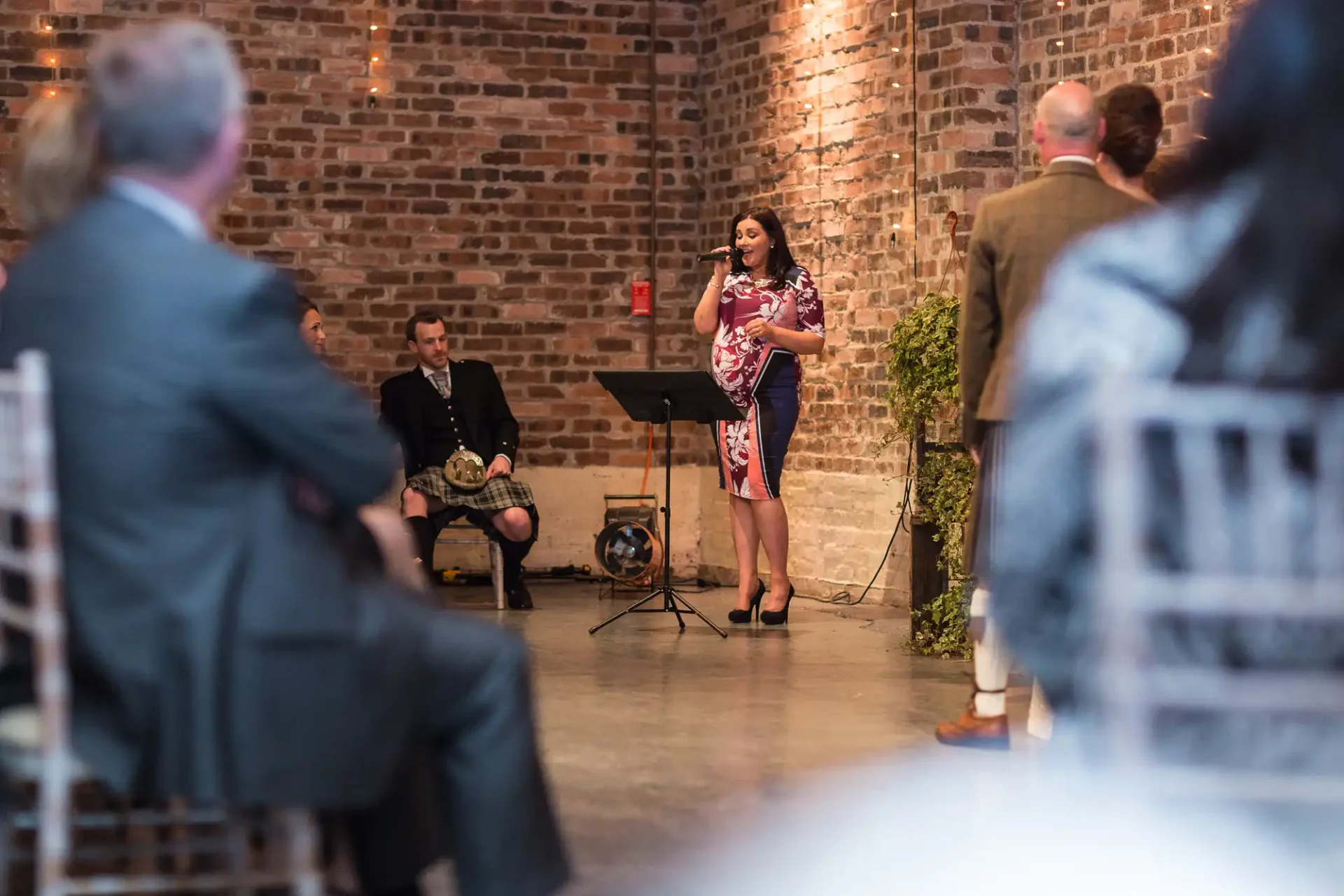 A woman gives a speech at a formal event in a brick-walled venue, with an audience listening attentively and a musician seated nearby.