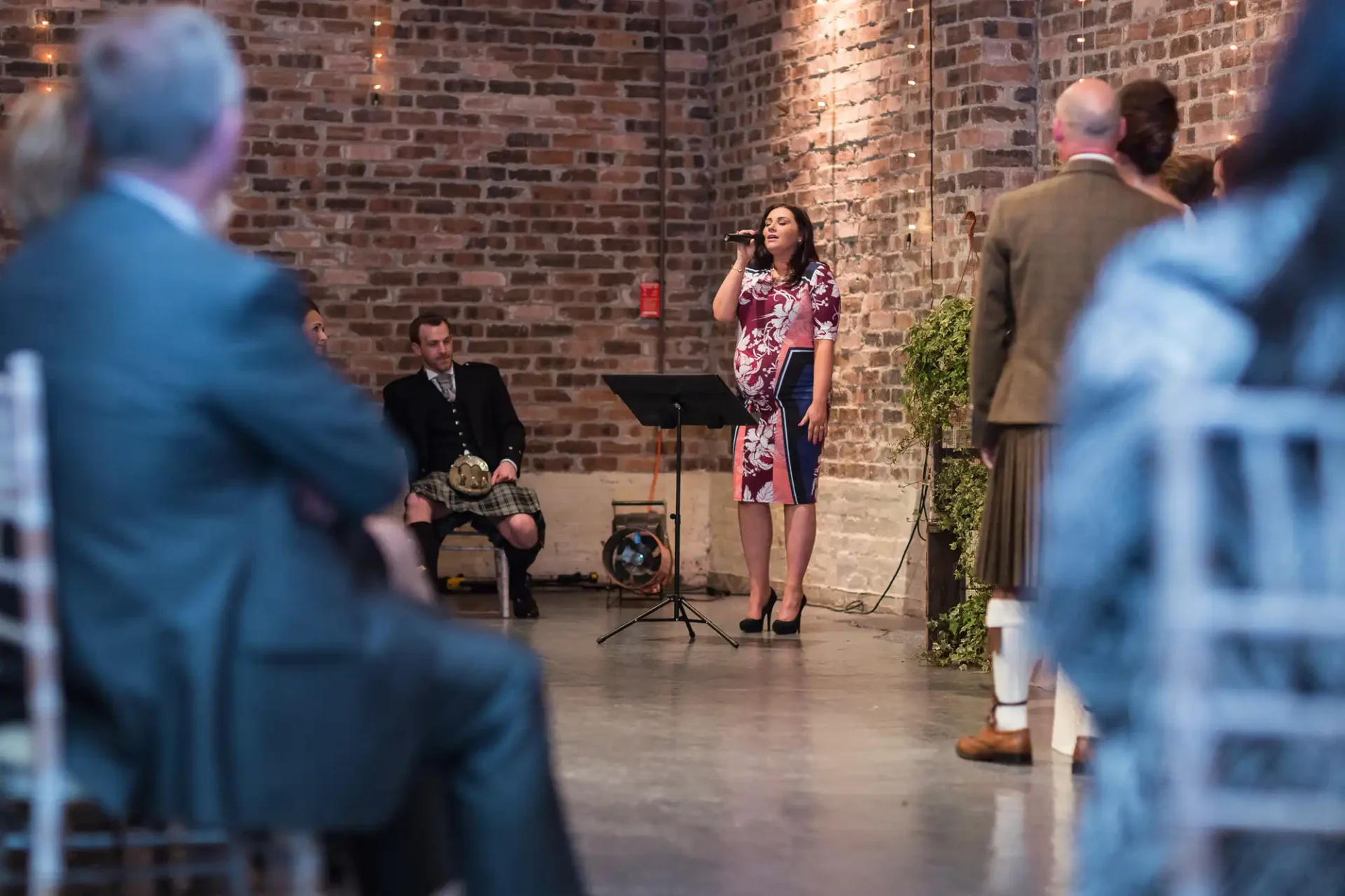 A woman singing into a microphone at a cozy event, with guests and a brick wall in the background, and a seated musician playing accordion.