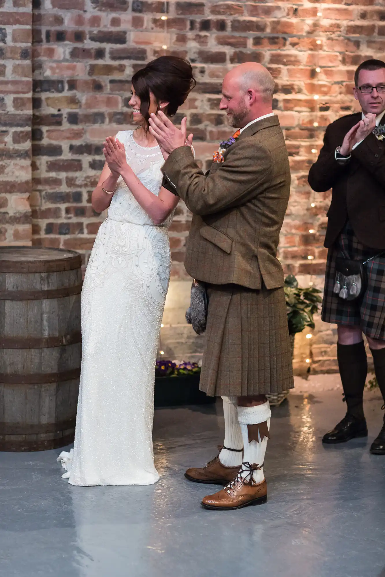 A bride and groom, the groom in a traditional kilt, embrace and admire the bride's ring during a wedding ceremony in a room with brick walls and barrels.
