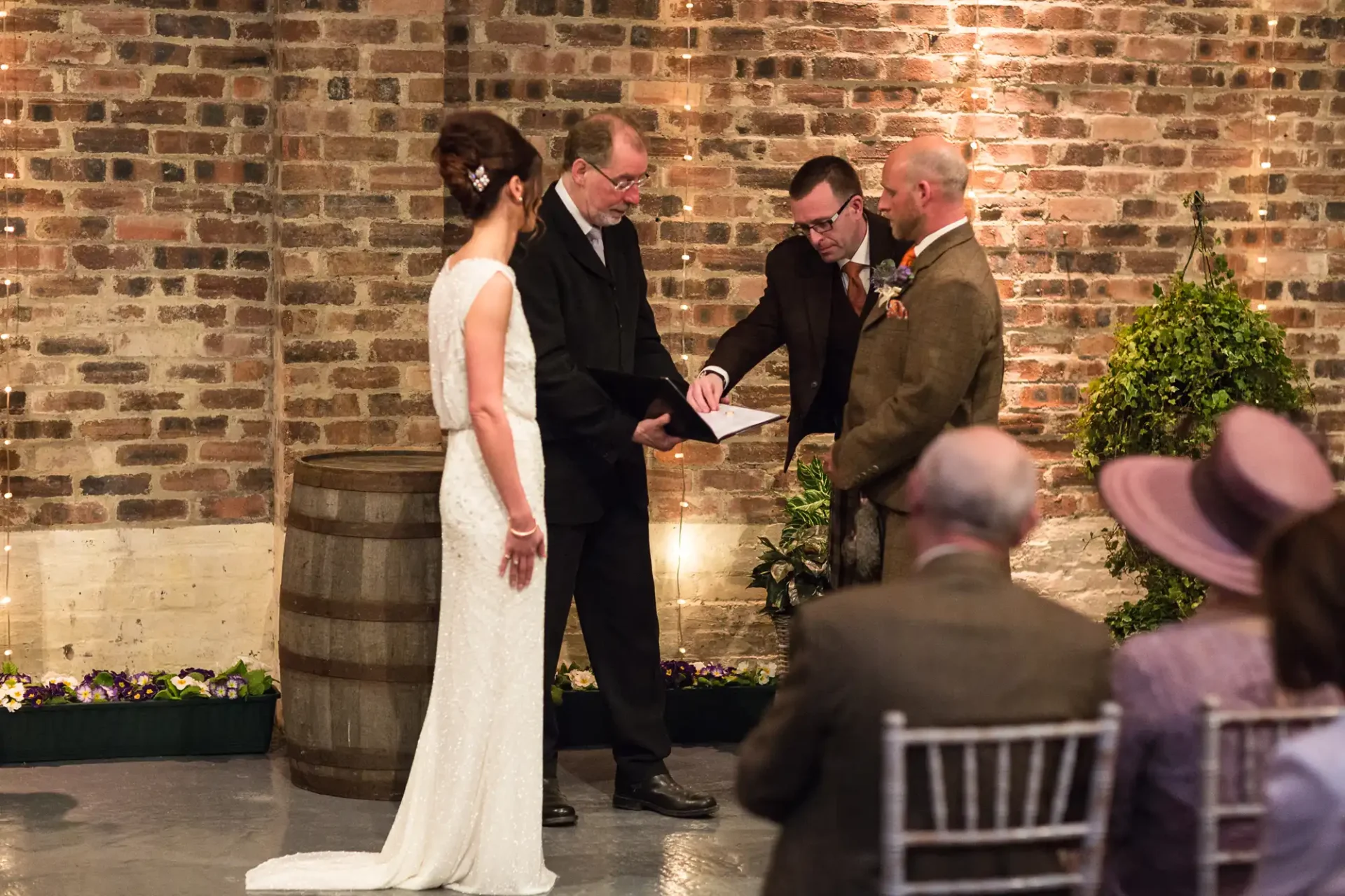 A wedding ceremony in progress inside a rustic venue with brick walls, featuring a bride and groom standing before an officiant, while two men look on. guests are seated observing.