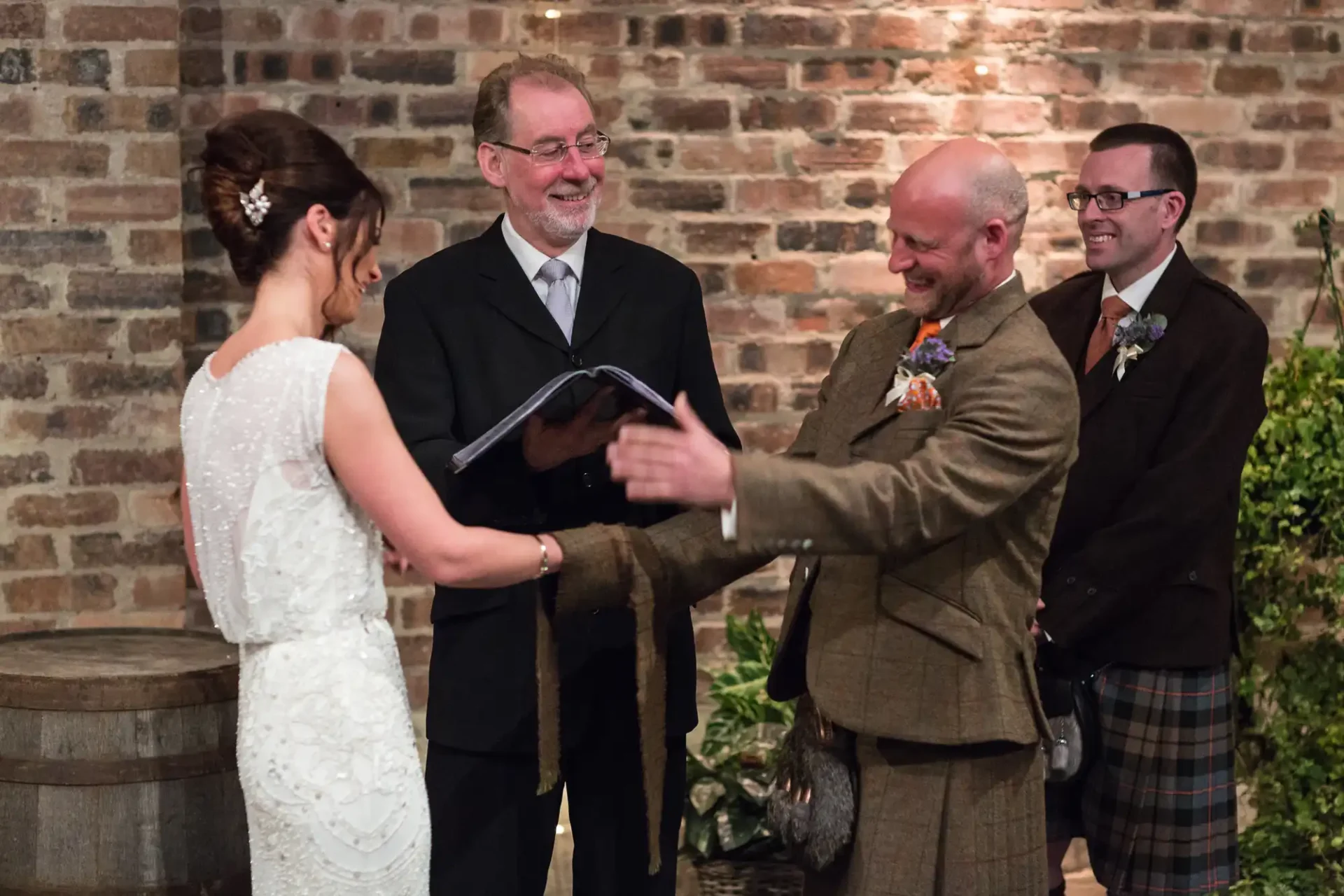 Bride and groom smiling at each other during their wedding ceremony, officiated by a man holding a book, with a guest smiling in the background.