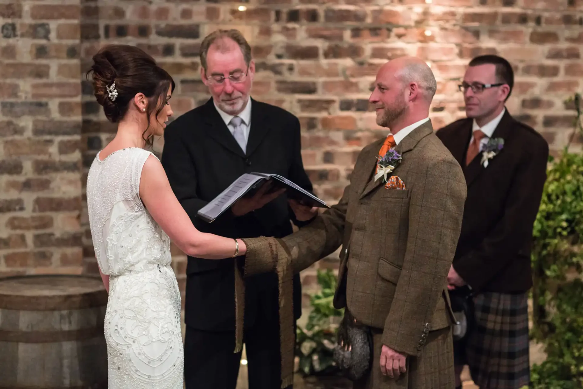 A bride and groom holding hands during their wedding ceremony, with an officiant and a best man standing beside them in a brick-walled venue.
