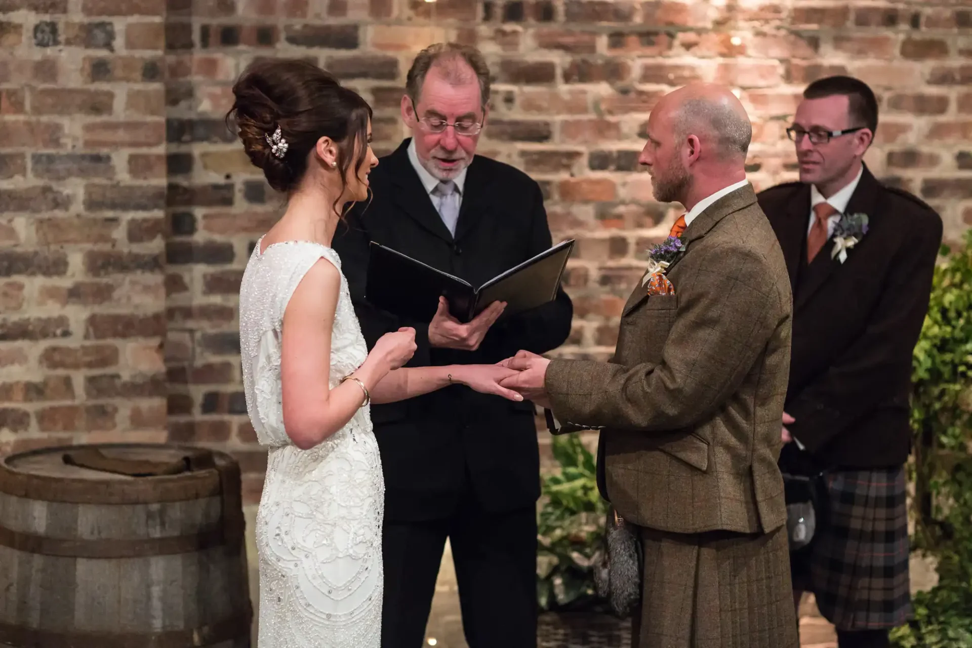 A wedding ceremony with a bride and groom exchanging vows, officiated by a clergyman, in a venue with rustic brick walls and barrel decor.
