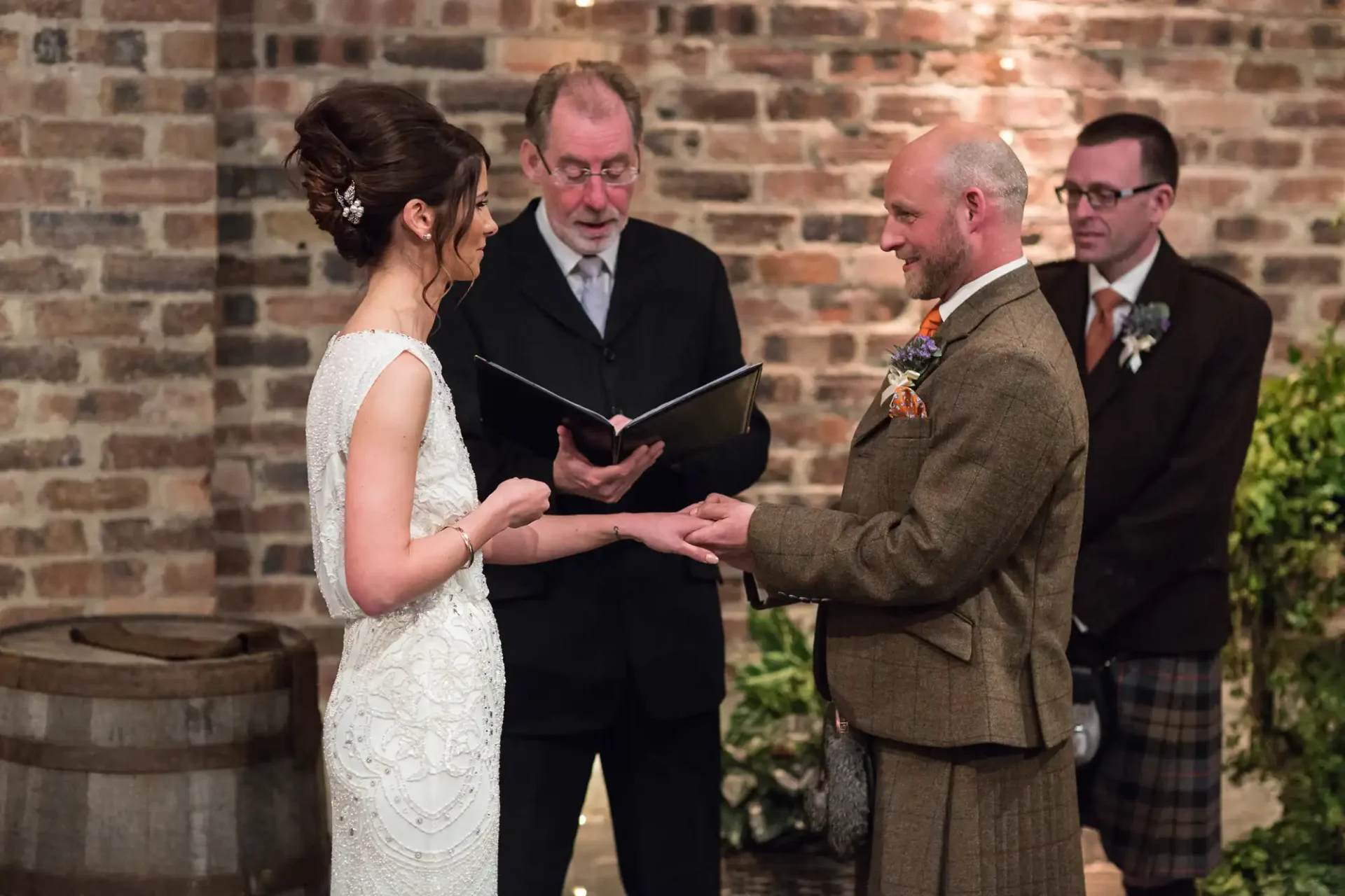A bride and groom exchange vows at an indoor wedding ceremony, officiated by a man holding a book, with another man observing in the background.