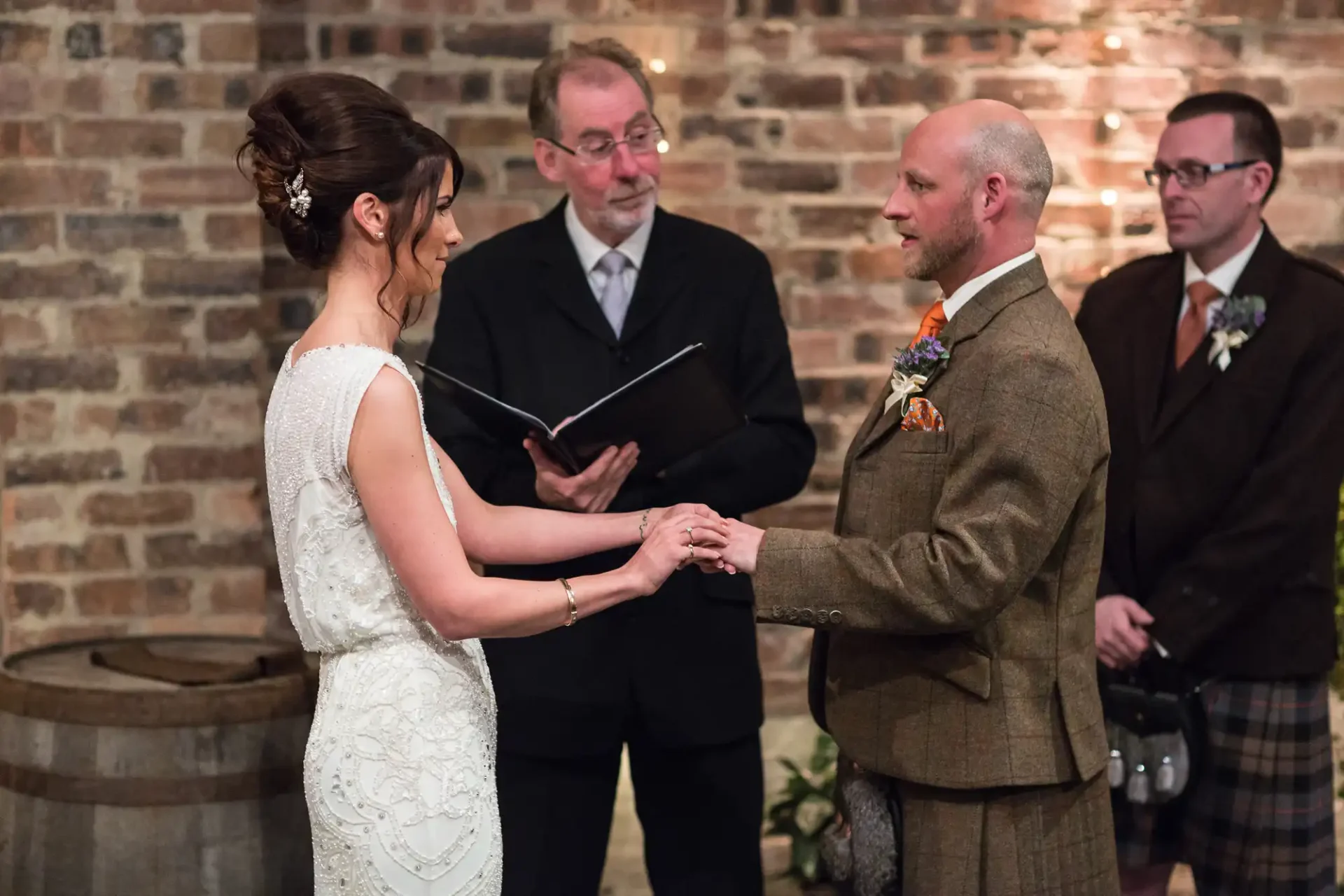 A bride and groom exchange vows, holding hands in front of an officiant at an indoor wedding ceremony with guests in the background.