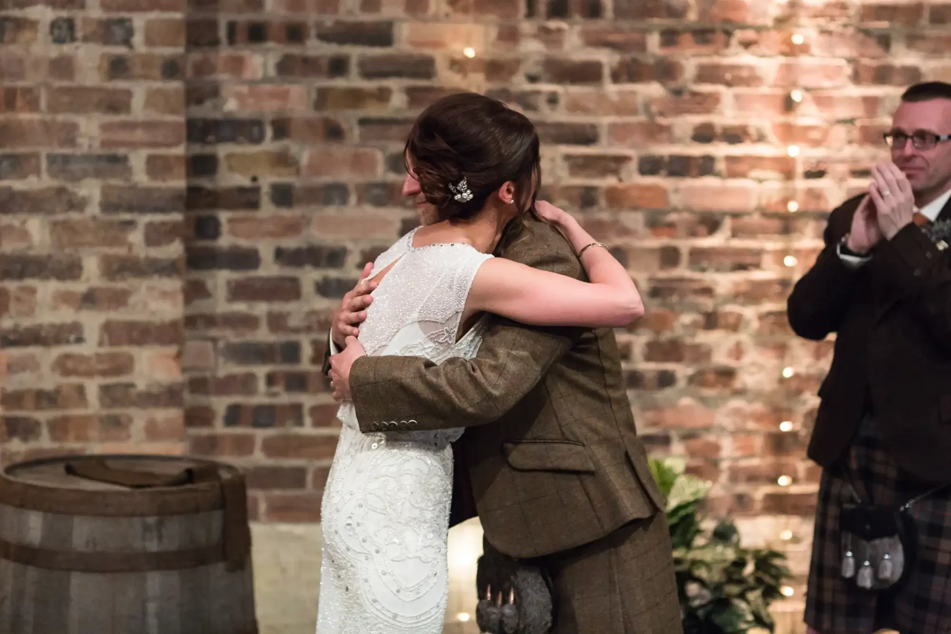 A bride and groom embrace lovingly during their wedding reception as a guest claps in the background, against a backdrop of brick walls and soft lights.
