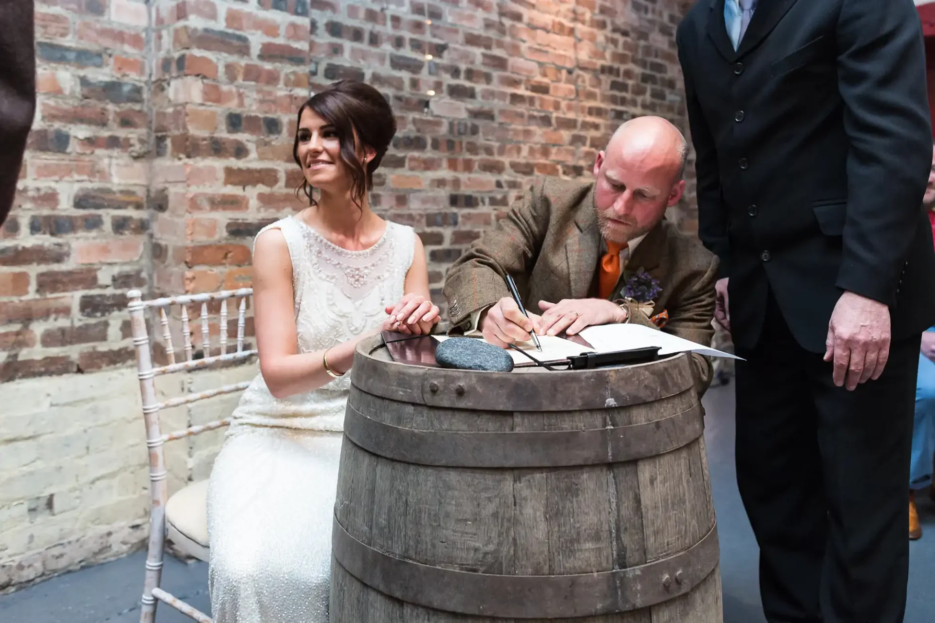 A bride and groom sign a document on a barrel in a rustic setting with brick walls, observing a wedding tradition.