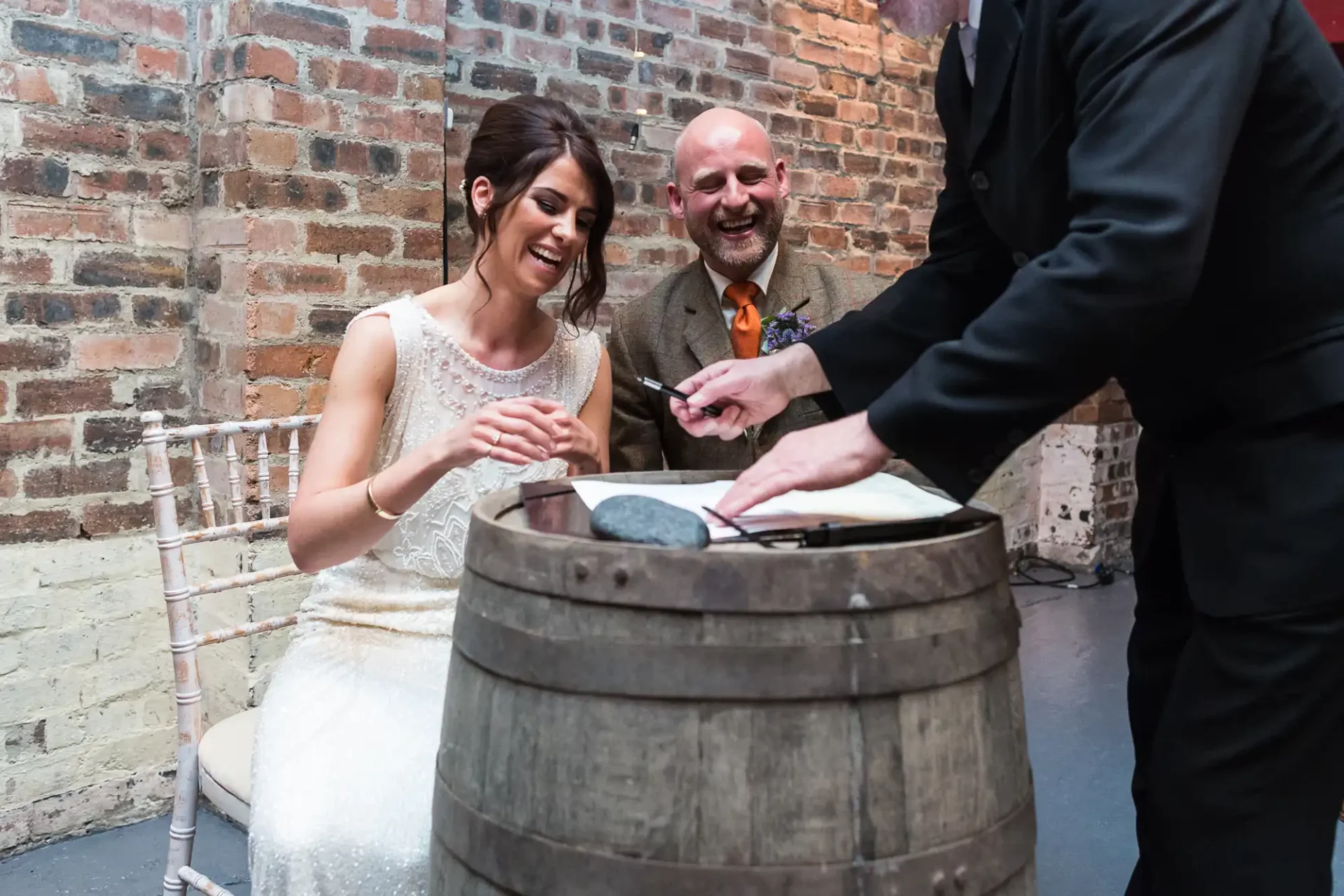 A bride and groom laugh joyfully while sitting and signing a document on a barrel, as an officiant stands beside them.
