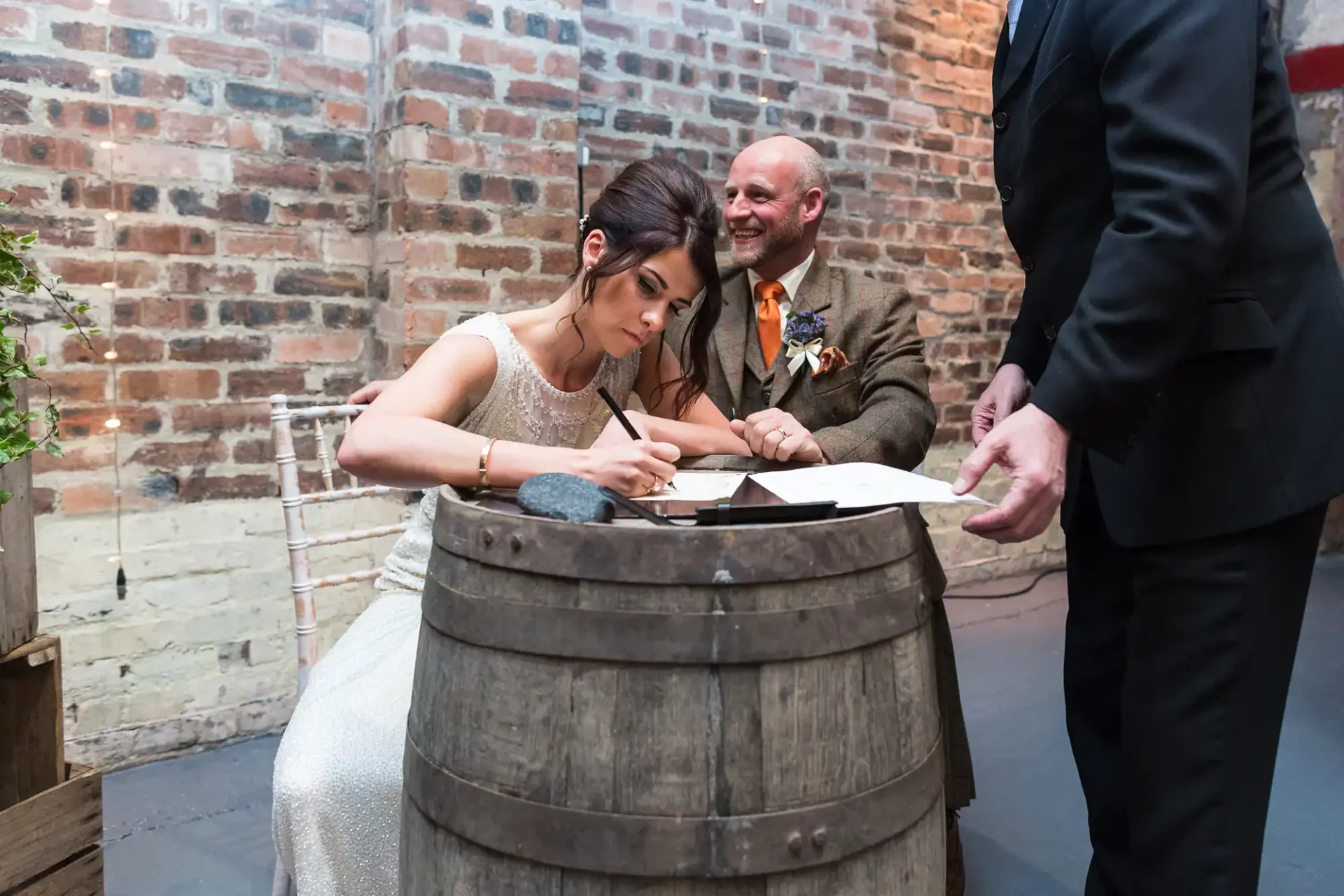 A bride and groom signing their marriage certificate on a barrel in a rustic brick-walled venue, with a man standing beside them.