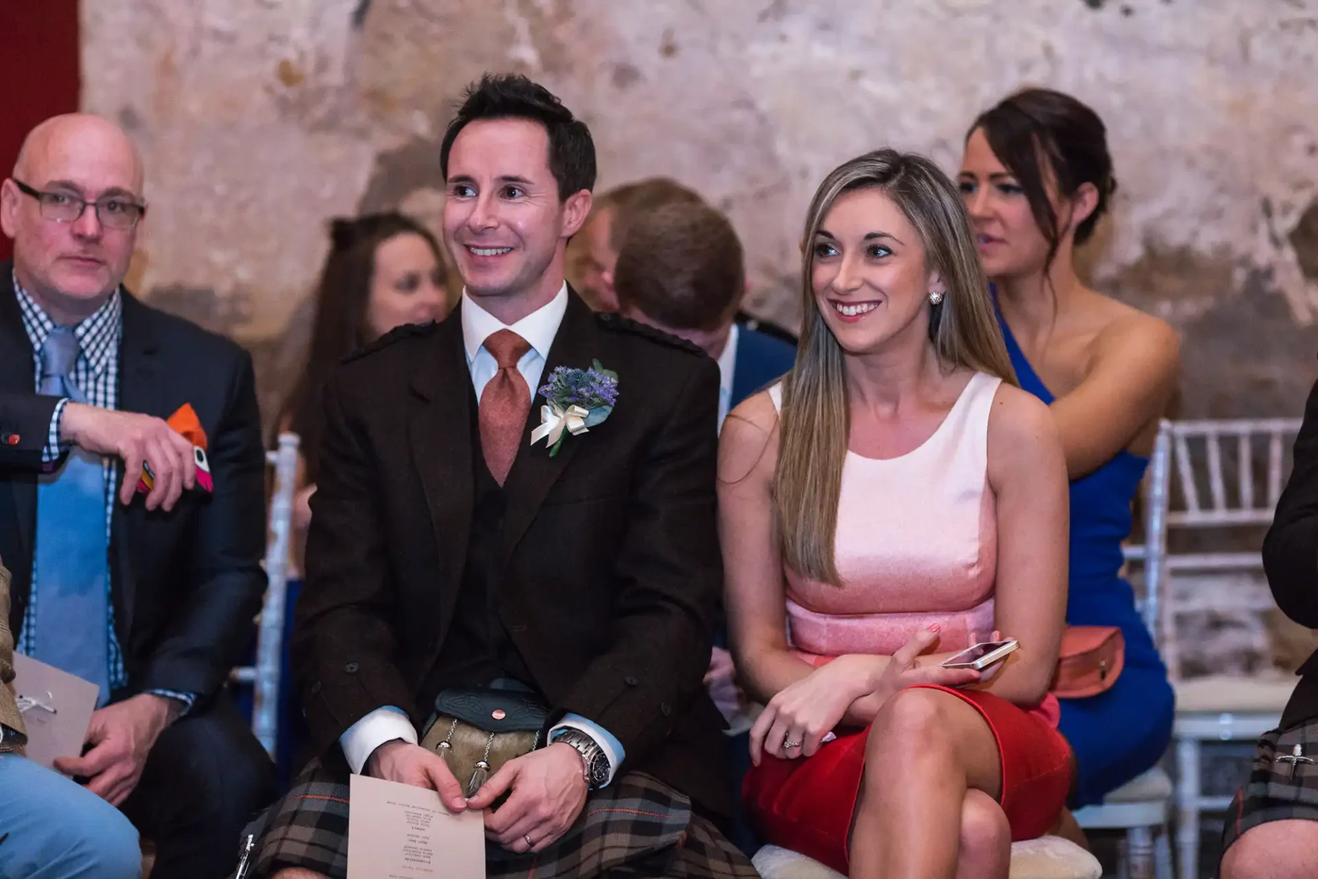 A man in a kilt and a woman in a pink dress sitting together at a wedding ceremony, both smiling and looking to the side.