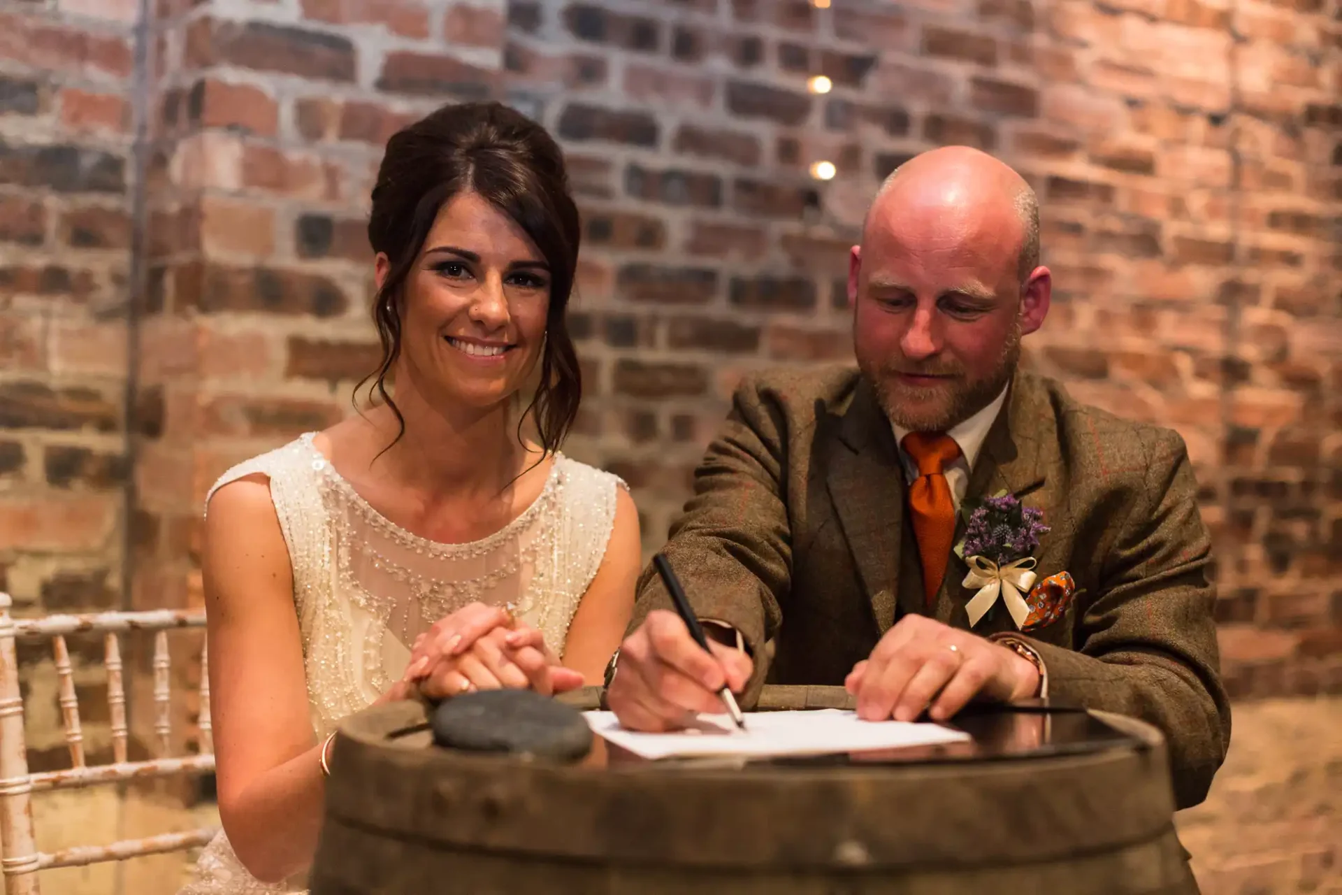 A bride and groom smiling while signing their marriage certificate at a table with a rustic brick backdrop.