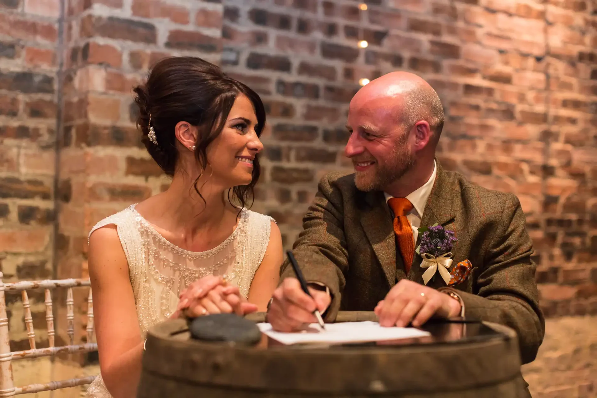 Bride and groom smiling at each other while signing a document, seated at a barrel table in a room with brick walls.