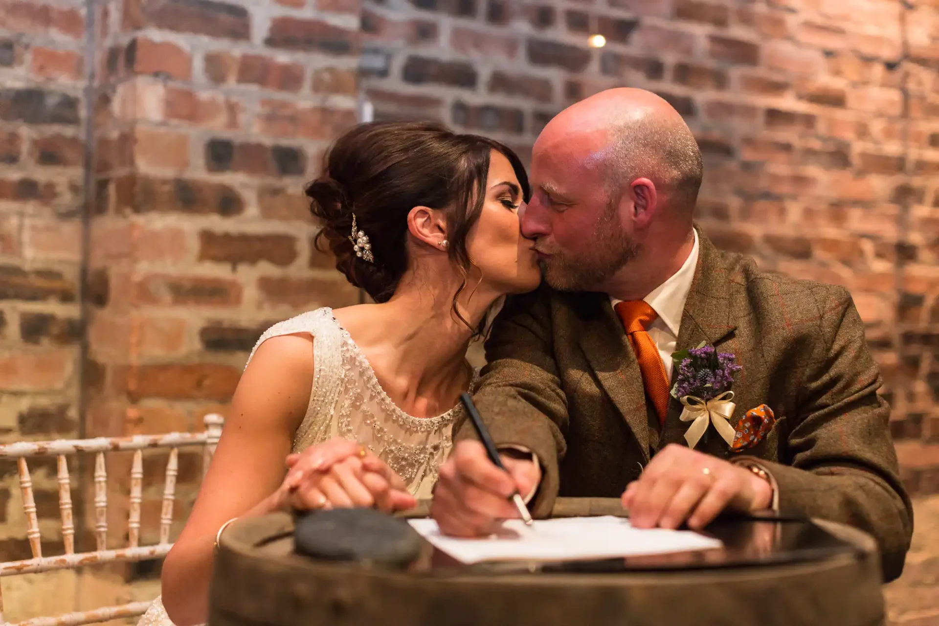 A bride and groom kiss while signing their marriage certificate at a table, in a warmly lit room with brick walls.