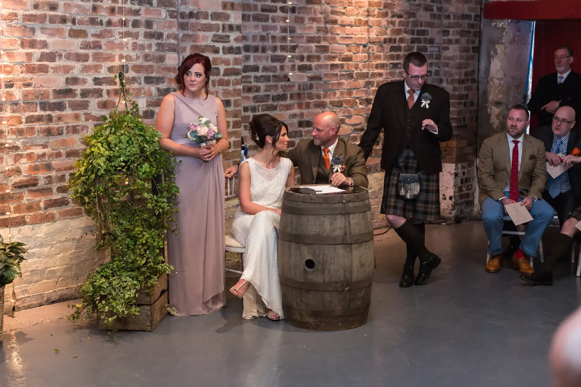 A wedding ceremony in a rustic venue with exposed brick walls, featuring a bride and groom seated by a wooden barrel, a bridesmaid standing nearby, and guests in varied attire watching.