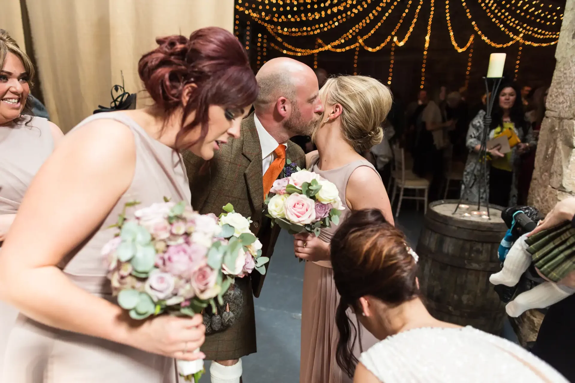 A bride and groom kiss while surrounded by bridesmaids at a warmly lit wedding venue with hanging string lights.