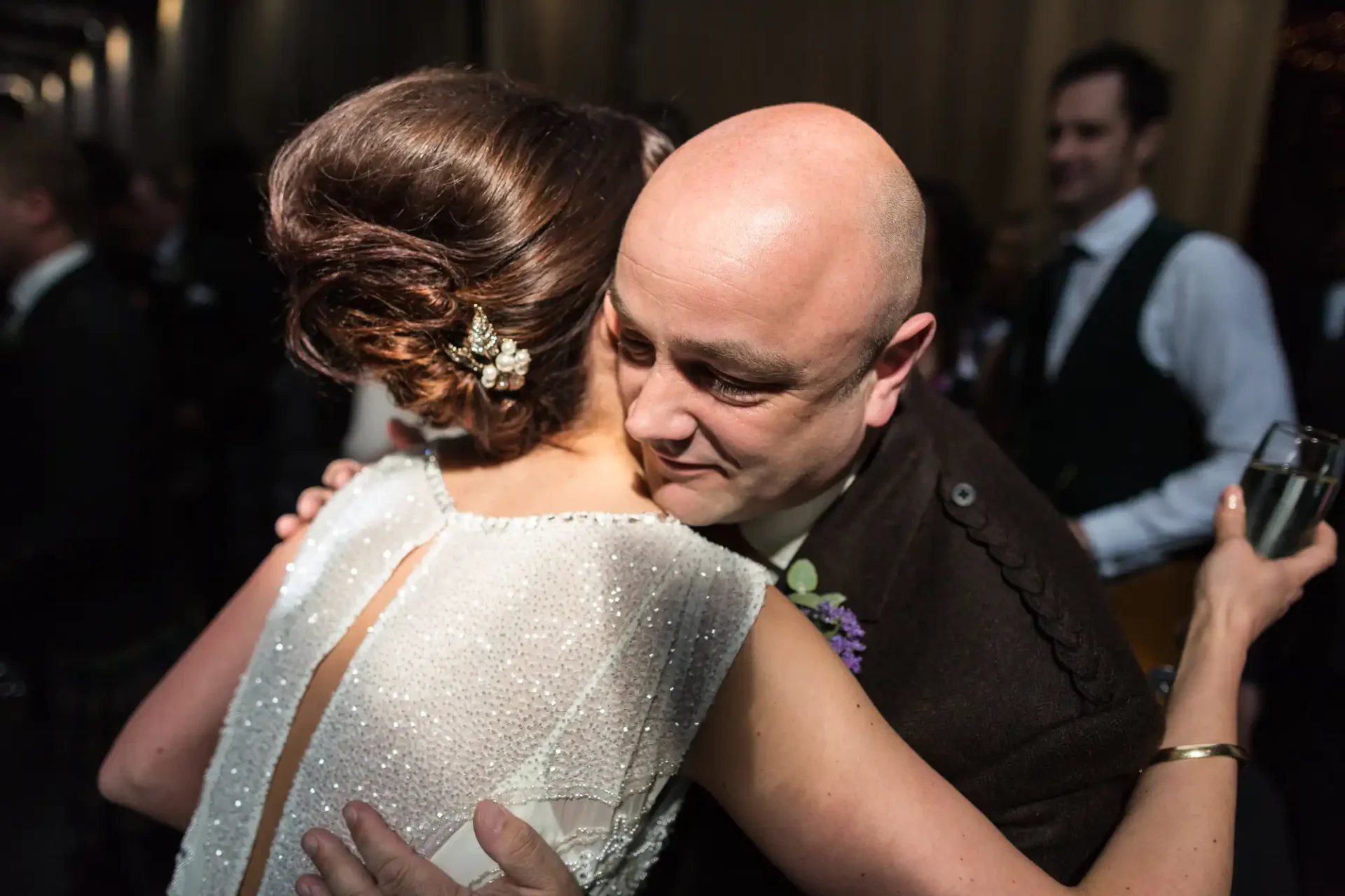 A man in a formal vest embraces a woman in a sequined wedding dress, both share a tender moment during a dimly lit reception.