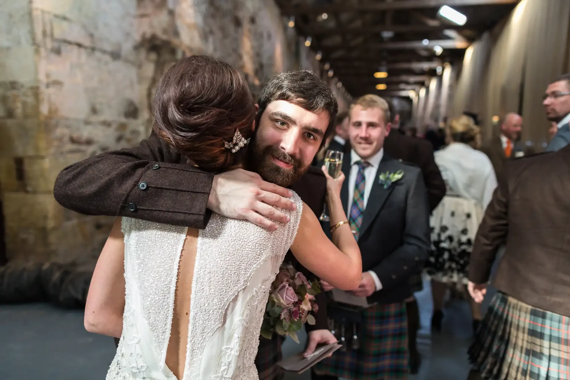 A bride embracing a man at a wedding reception, with a man in a kilt holding a glass of wine in the background.